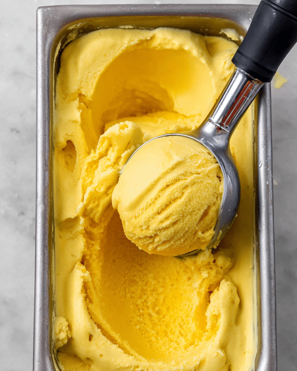 The image shows a silver metal container filled with bright yellow, smooth and creamy ice cream. The ice cream has a thick, soft texture with some small ice crystals on the surface. A shiny metal ice cream scoop with a black handle is in the process of lifting a round scoop of the ice cream from the container, showing the creamy inside. The container is resting on a white marbled texture surface. photo taken with an iphone --ar 4:5 --v 7