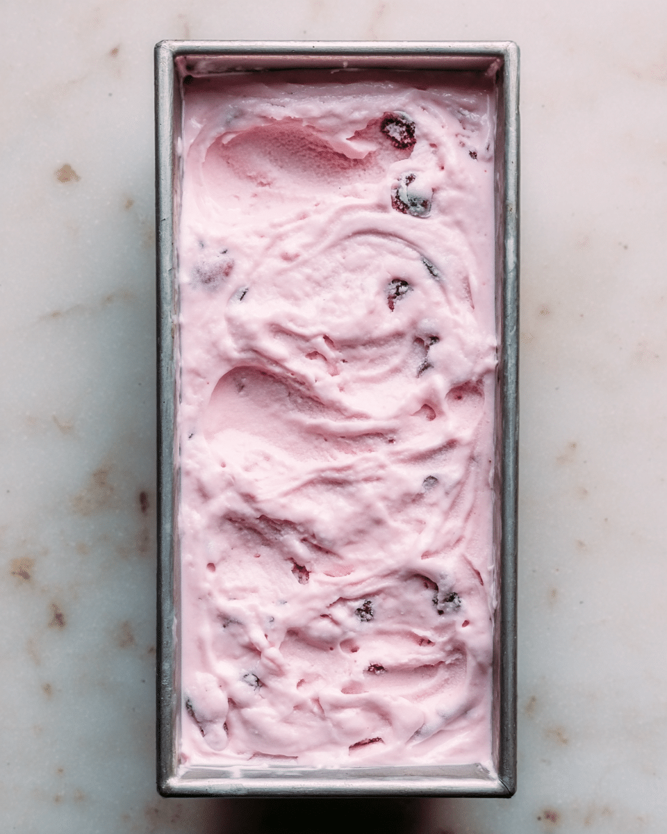 The image shows a rectangular metal container filled with a layer of light pink frozen dessert, which has a soft, creamy texture with small darker pink and purple spots spread throughout. The surface is uneven with swirled and slightly bumpy areas, giving a fresh and rich look. The container sits on a white marbled surface. Photo taken with an iphone --ar 4:5 --v 7
