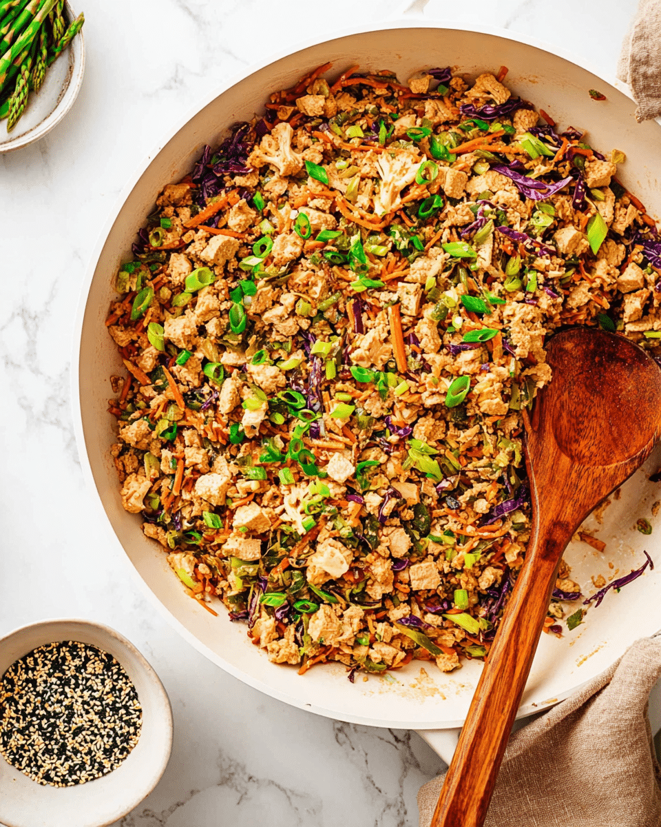 A large white pan filled with a stir fry mix that has finely chopped light brown tofu pieces as the main layer, mixed with thin strips of orange carrots and purple cabbage, and small bits of light beige cauliflower scattered throughout. On top, bright green chopped scallions are sprinkled evenly, adding fresh color. A wooden spoon with visible grain rests inside the pan, partially submerged in the mix. The pan sits on a white marbled surface with a light brown cloth near its handle, and next to it, a small white bowl with mixed black and white sesame seeds and seaweed flakes, and a partial view of a white bowl with green asparagus spears. photo taken with an iphone --ar 4:5 --v 7