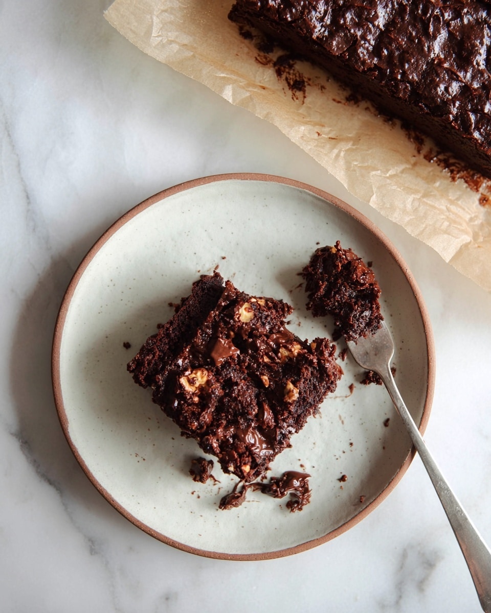 A single square piece of dark chocolate brownie sits on a white plate with a slight brown rim, showing a rich, textured top with chunks of nuts or chocolate pieces embedded. The brownie has a dense, crumbly interior with some crumbs scattered around it on the plate. A silver fork lies on the right side, holding a small broken piece of the brownie, with a bit of chocolate spread visible near the crumbs. The plate rests on a white marbled surface, and above it, a larger uncut brownie block is partially visible on parchment paper. photo taken with an iphone --ar 4:5 --v 7