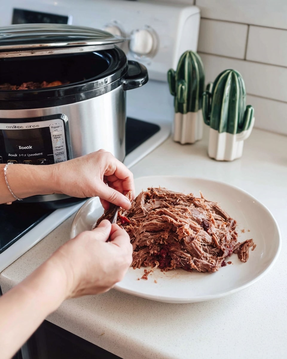 A slow cooker filled with dark brown cooked meat sits on a white marbled countertop next to a white stove. In the foreground, a woman's hands are shredding a large piece of the cooked meat on a plain white round plate. The meat has a tender texture with visible strands being pulled apart. Behind the cooker, there are two small decorative green and white ceramic cactus figures. The scene is bright and clean with soft natural light. Photo taken with an iphone --ar 4:5 --v 7