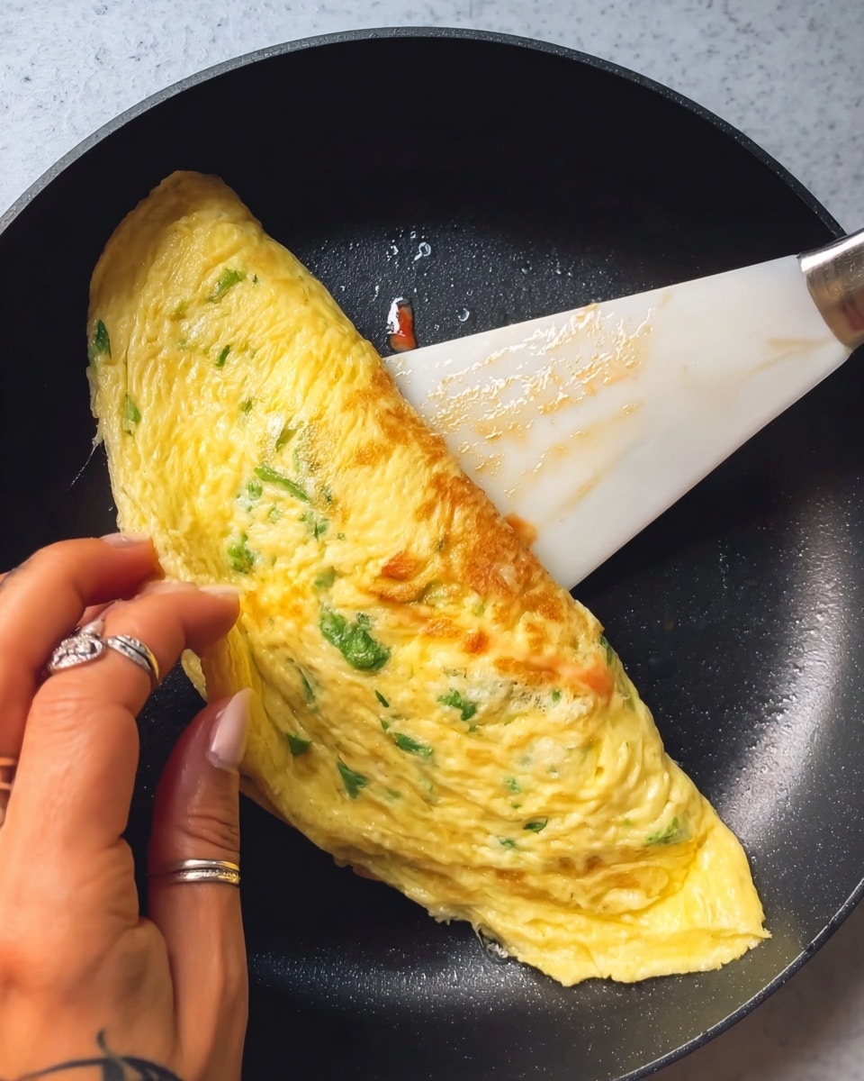 The image shows a close-up of a soft, yellow omelette being folded in a black frying pan with a white spatula on the right side. The omelette has visible green herbs mixed inside and some melted cheese or sauce lightly oozing out from the bottom edge. A woman's hand with rings and a small tattoo is gently lifting one side of the omelette to fold it over. The pan surface is black but described as a white marbled texture for the prompt. Photo taken with an iphone --ar 4:5 --v 7