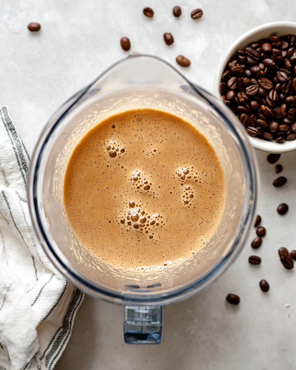 A top view of a clear blender jar filled with a creamy, light brown mixture that has small bubbles on its surface. The blender jar is placed on a white marbled texture surface. To the upper right of the jar, there is a small white bowl filled with dark brown roasted coffee beans, and scattered coffee beans can be seen on the surface nearby. A white kitchen towel with black stripes is partially visible on the left side near the jar. photo taken with an iphone --ar 4:5 --v 7