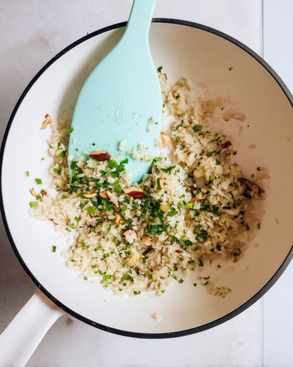 A white pan with a black edge holds a small pile of light beige cooked rice mixed with green chopped herbs and small pieces of nuts or seeds. A light blue spatula rests inside the pan, holding some of the rice mixture. The background is a white marbled surface. photo taken with an iphone --ar 4:5 --v 7