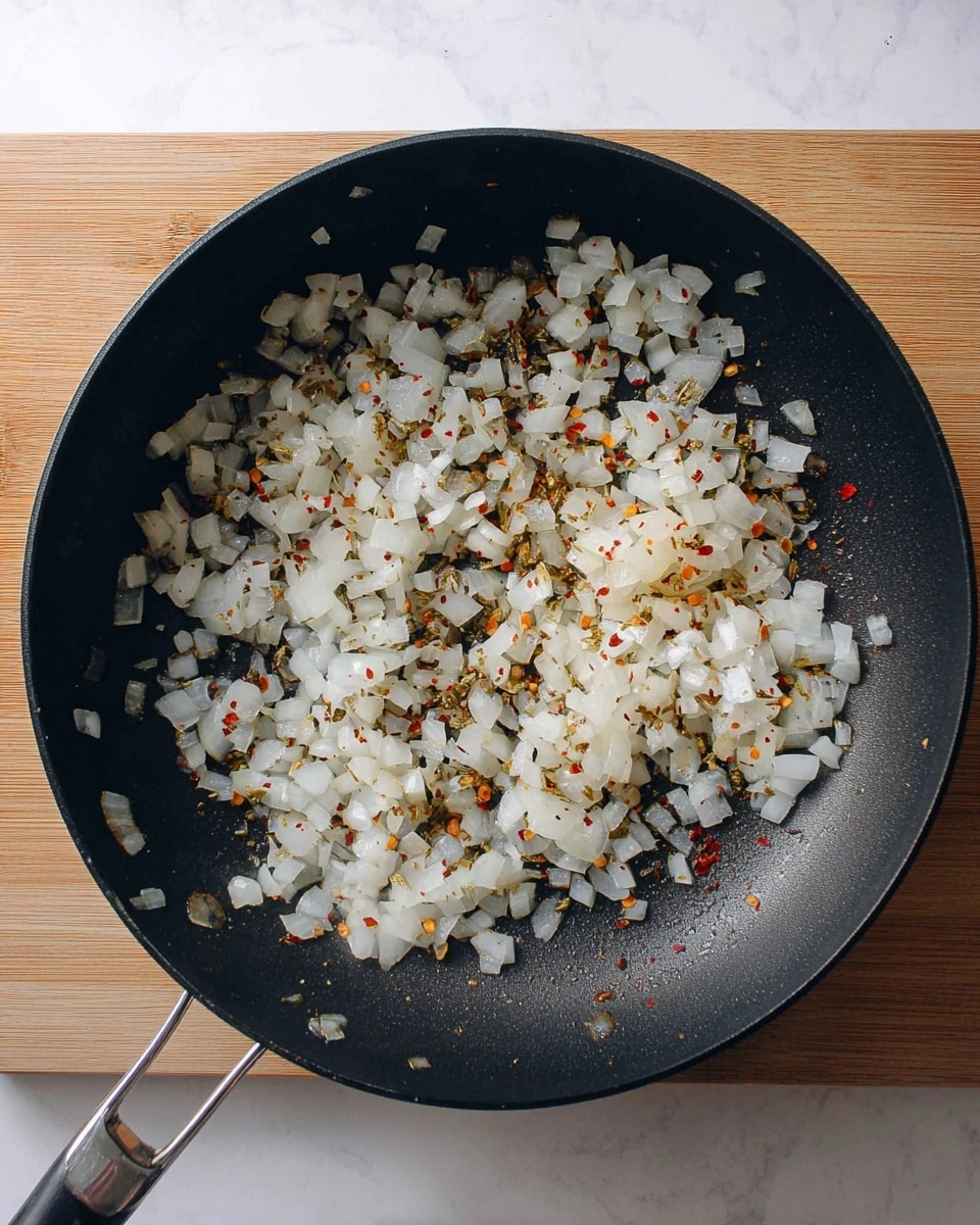 A black frying pan filled with one layer of diced white onions that have been cooked until translucent, mixed evenly with small bits of red pepper flakes and dried herbs, giving a speckled texture of red and green on the onions; the pan rests on a light wood surface replaced by a white marbled texture background in the image, viewed from above showing the full round pan and its metal handle on the left side, with the cooked onions spread mostly around the pan's center and edges, leaving some empty black space visible; photo taken with an iphone --ar 4:5 --v 7