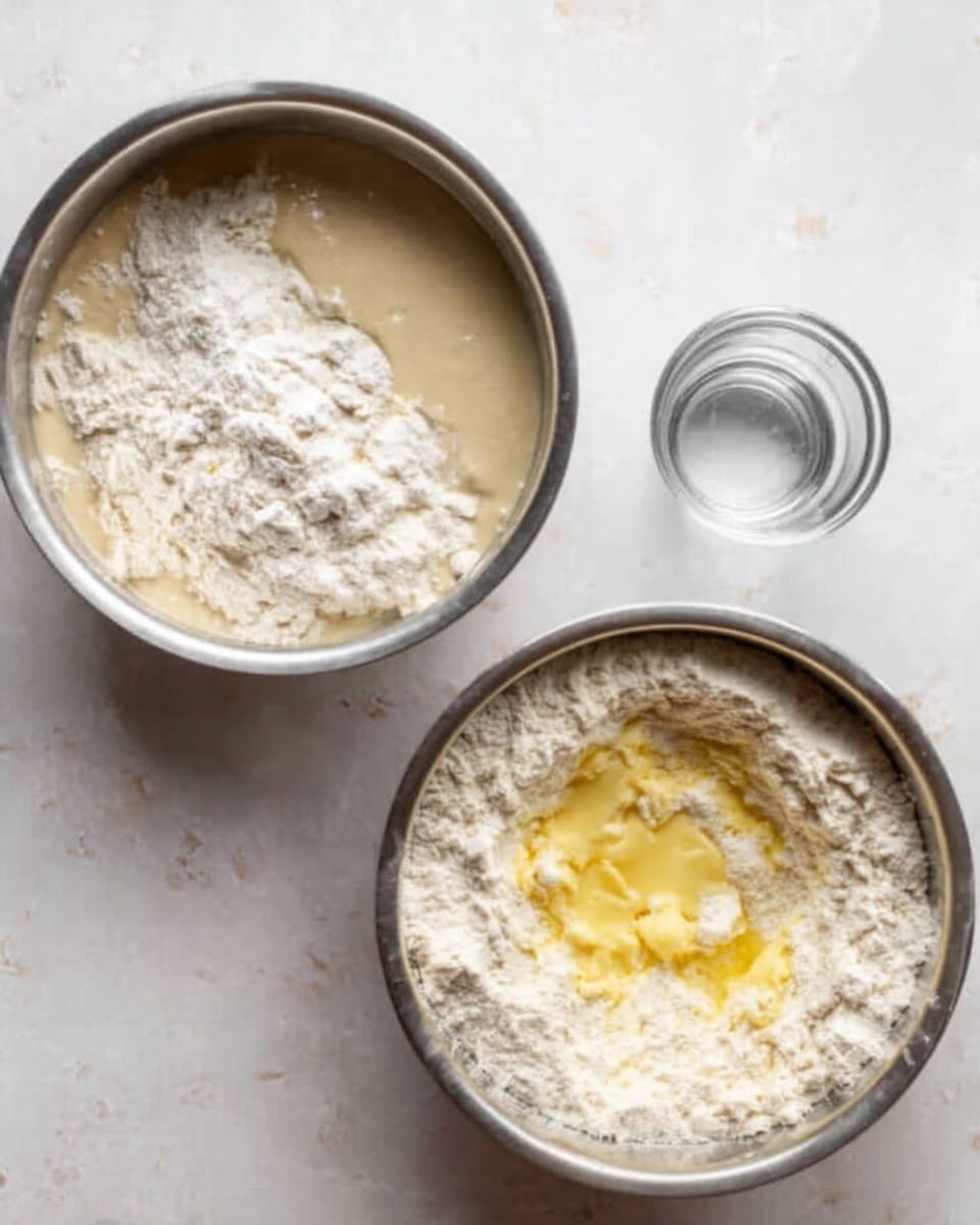 The image shows two round metal bowls on a white marbled textured surface. The left bowl contains a mix of white flour and a beige liquid, which has a smooth texture and is partly absorbed by the flour around it. Next to this bowl is a small transparent glass with a bit of clear liquid inside. The right bowl also has white flour but with a layer of melted yellow butter on top, surrounded by the flour that holds it like a ring. The texture contrasts between the powdery flour and the oily butter are clear. photo taken with an iphone --ar 4:5 --v 7