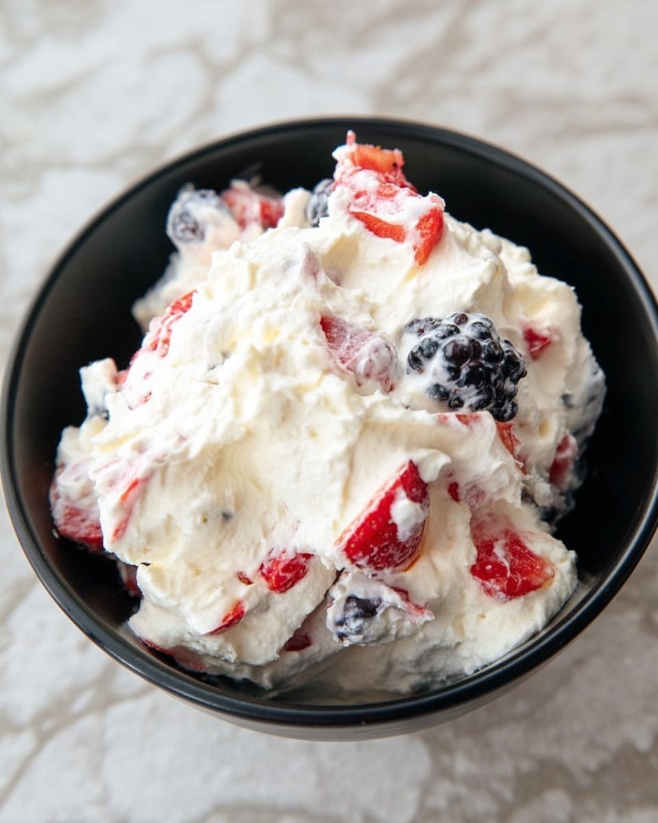 A black bowl filled with a fluffy mix of white whipped cream and pieces of red strawberries and dark blue-black blackberries. The cream looks soft and smooth, holding the bright fruit pieces mixed evenly throughout. The bowl sits on a white marbled surface, showing the contrast between the dark bowl and the colorful creamy mixture inside. photo taken with an iphone --ar 4:5 --v 7