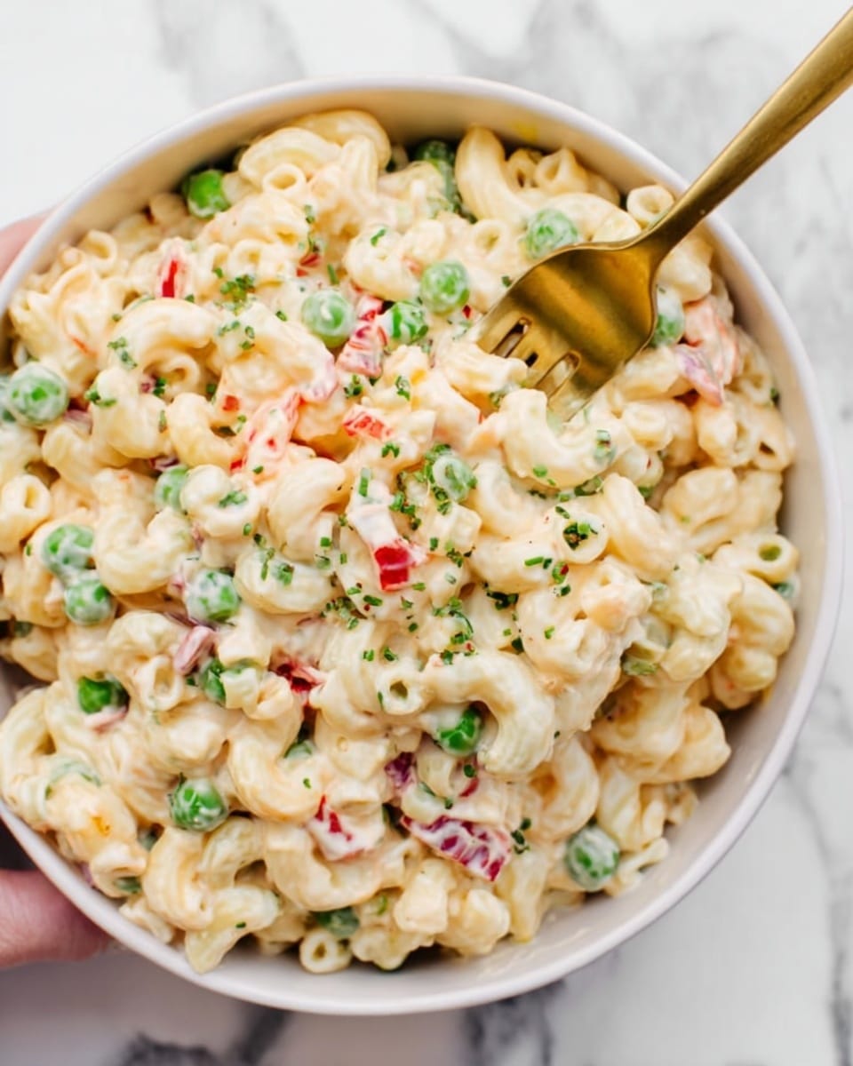 The image shows a white bowl full of creamy macaroni salad on a white marbled background. The macaroni is light yellow and coated with a thick creamy dressing. There are green peas and small pieces of red bell pepper mixed evenly throughout. A woman's hand is holding a gold fork, digging into the salad from the right side of the bowl. The salad looks fresh with small bits of green herbs sprinkled on top. Photo taken with an iphone --ar 4:5 --v 7