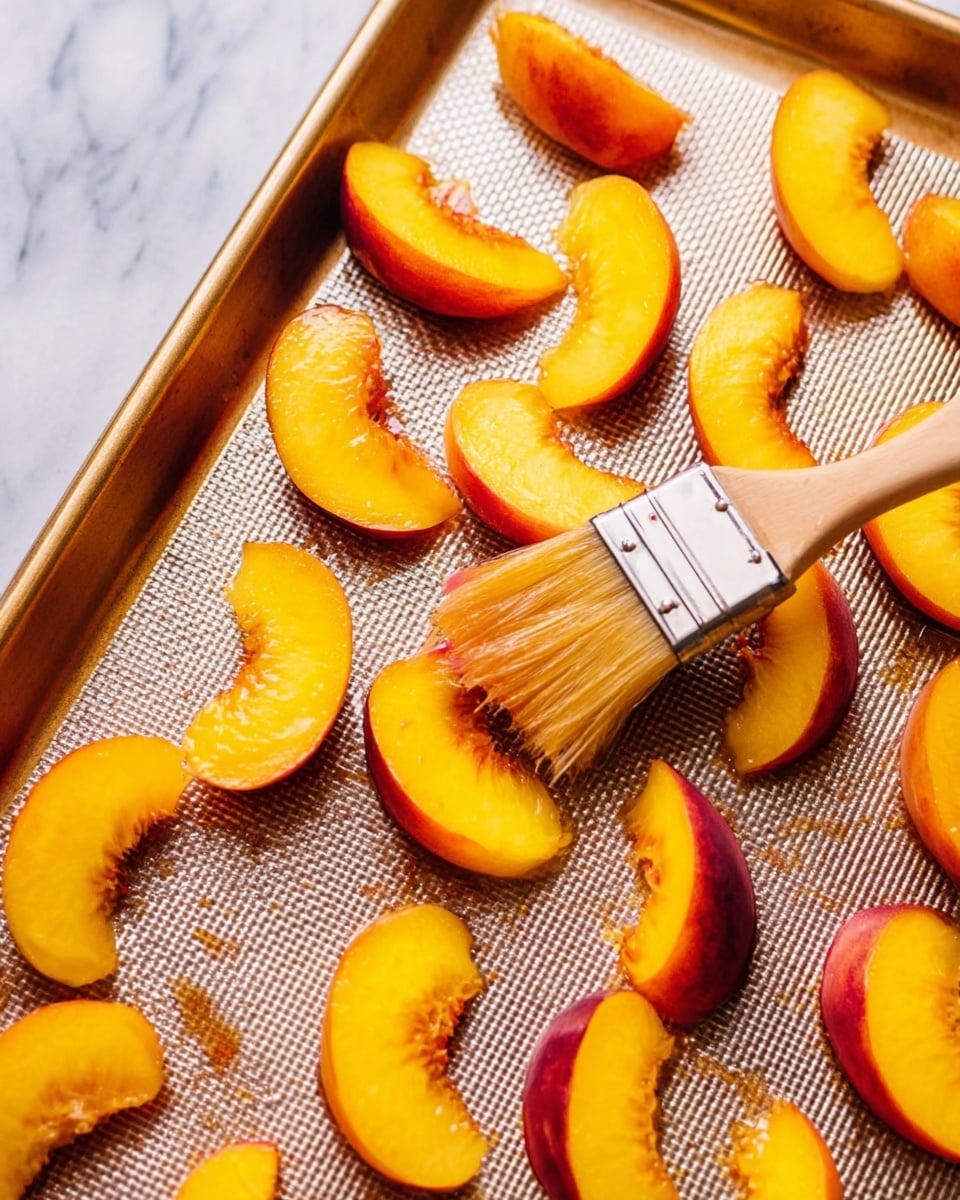 The image shows a baking tray with a textured surface holding many peach slices arranged in rows. Each slice has a bright yellow flesh with a red-orange skin edge, and a glossy brush with light brown bristles is spreading a clear liquid on one slice near the center. The baking tray is metal and sits on a white marbled surface. The overall look is fresh and vibrant, highlighting the juicy texture of the fruit. photo taken with an iphone --ar 4:5 --v 7
