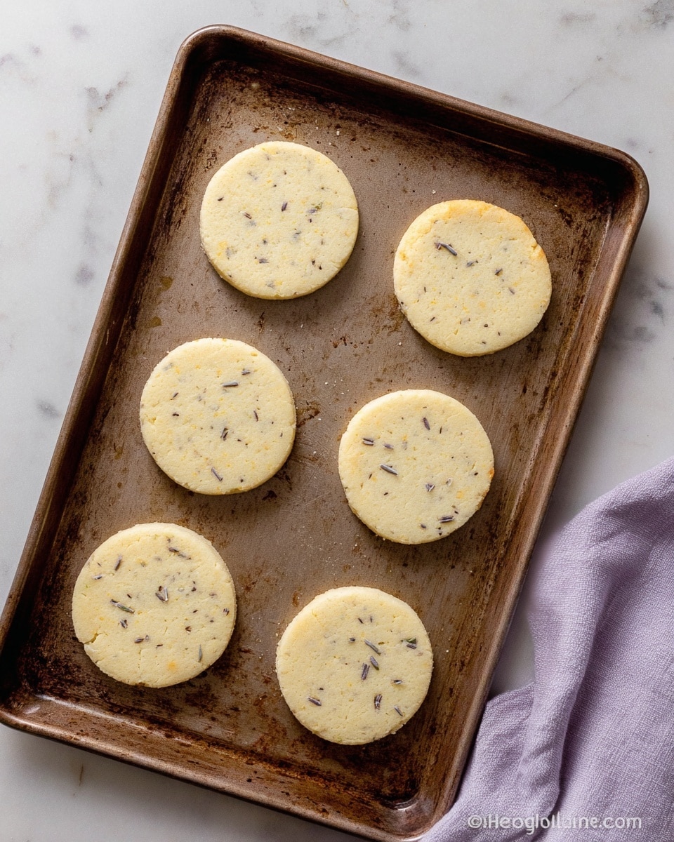 The image shows six round, pale yellow cookies on a brown baking tray with some darker oily spots around them. The cookies have small dark specks mixed evenly throughout and are spaced apart in two rows. To the right side of the tray, there is a light purple cloth partially visible. The tray is placed on a white marbled surface. The lighting is soft, making the cookies look fresh and ready to bake or just baked. photo taken with an iphone --ar 4:5 --v 7