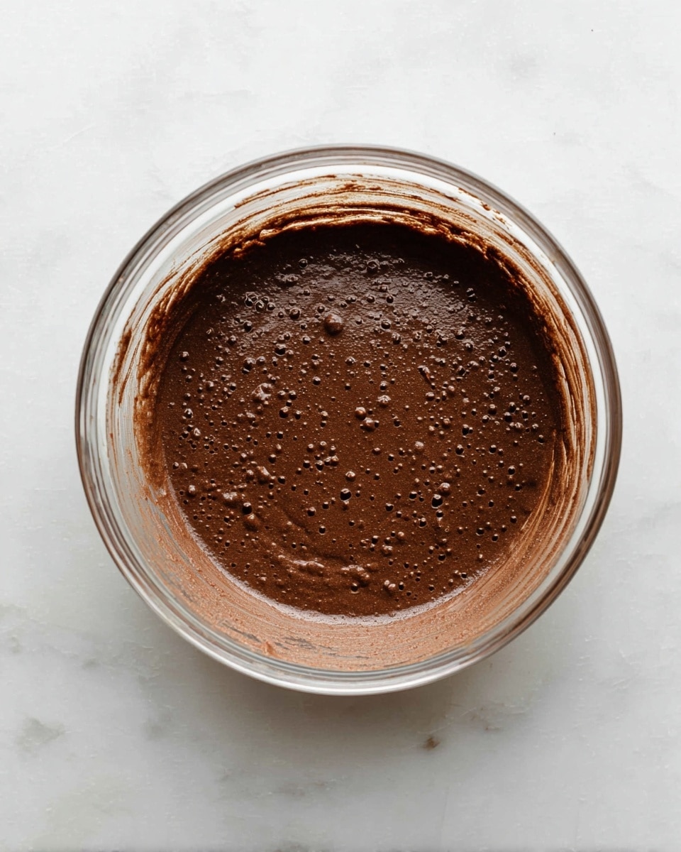 A clear glass bowl filled with a thick, dark brown chocolate batter that has small bubbles on its smooth surface. The batter spreads evenly inside the bowl, leaving some traces along the inner sides of the bowl. The bowl rests on a white marbled surface. photo taken with an iphone --ar 4:5 --v 7