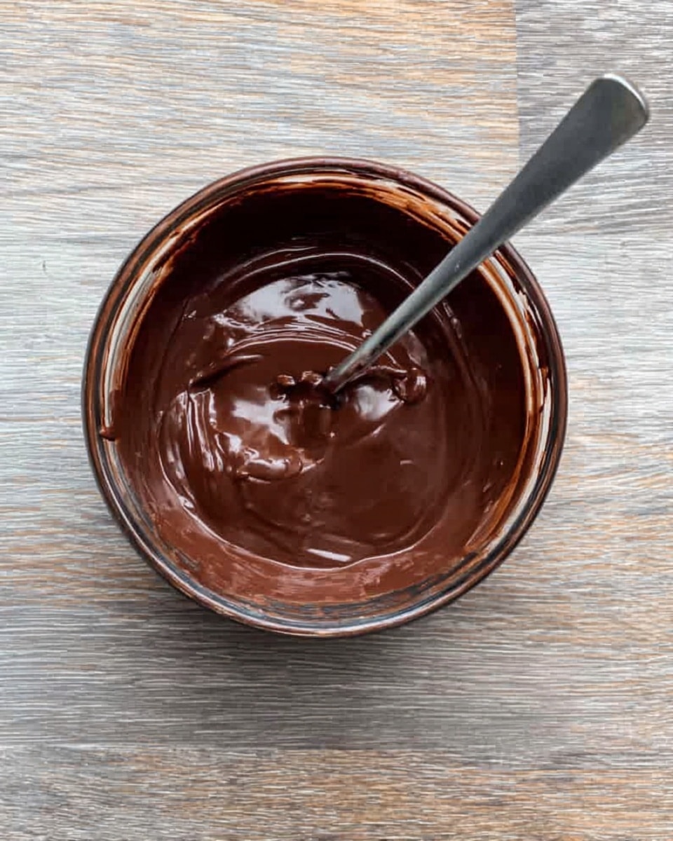A clear glass bowl filled with thick, smooth dark brown chocolate spread with a shiny surface, a silver spoon resting inside the bowl spreading the chocolate, all placed on a white marbled surface with a wooden texture background visible around the bowl’s edges, photo taken with an iphone --ar 4:5 --v 7