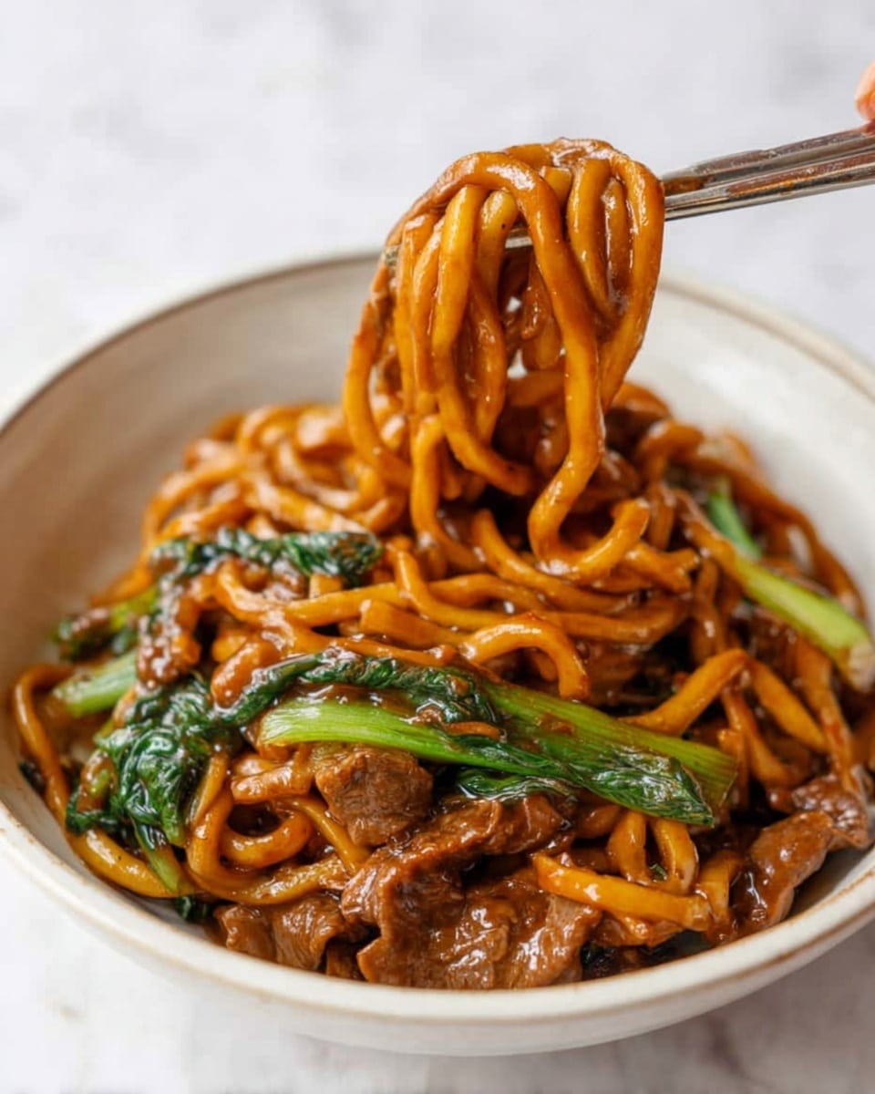 A white bowl filled with thick, shiny noodles coated in a dark brown sauce. The noodles are mixed with pieces of cooked meat, green leafy vegetables, and some green stems. A pair of metal chopsticks held by a woman's hand lifts a thick bundle of glossy noodles above the bowl. The background and surface show a white marbled texture. photo taken with an iphone --ar 4:5 --v 7
