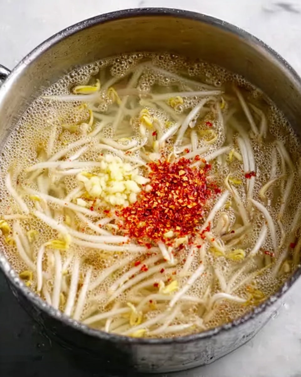 A close-up view of a metal pot filled with boiling broth and cooked bean sprouts, which are pale yellow and white in color. The broth looks clear with floating small bubbles, giving a light texture. On top of the sprouts, there's a small pile of finely chopped garlic in the center, surrounded by a sprinkle of bright red chili flakes. The pot is set against a white marbled surface. photo taken with an iphone --ar 4:5 --v 7