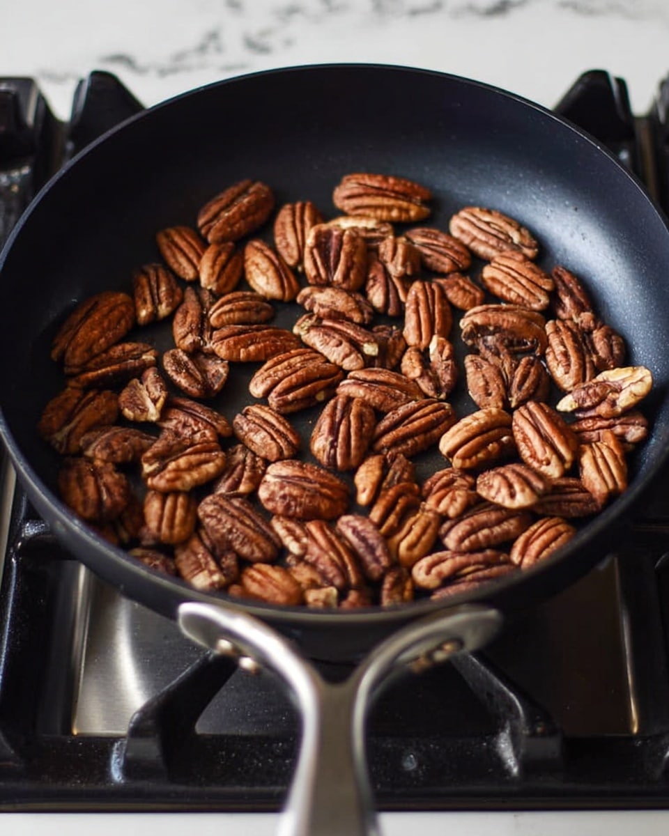 A black frying pan filled with whole brown pecan nuts spread evenly inside, showing their rough textured shells with lighter ridges. The pan rests on a black stove burner with silver metal handles, and the background is a white marbled surface. The pecans are warm and toasting, giving a cozy cooking feeling photo taken with an iphone --ar 4:5 --v 7