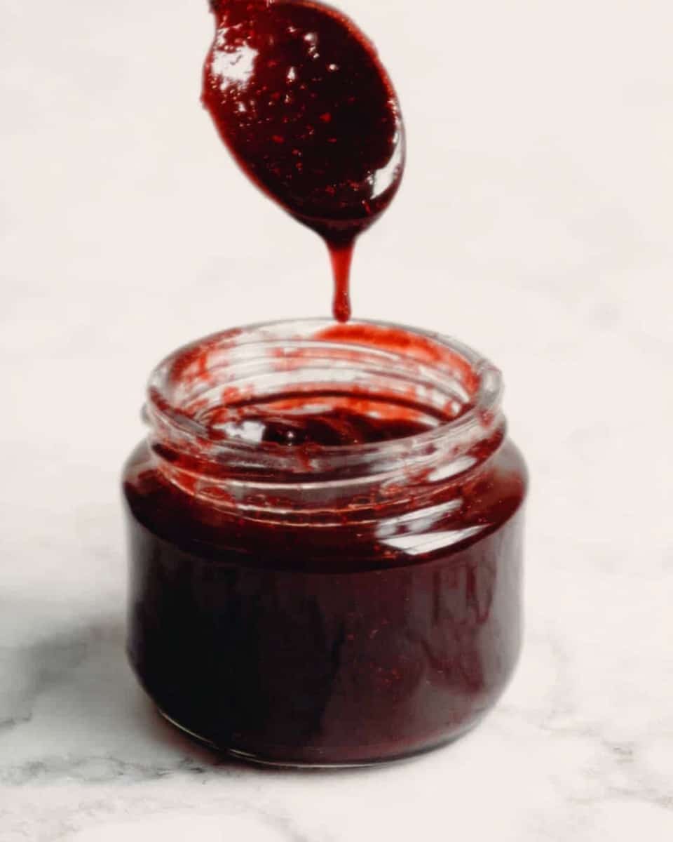 A small clear glass jar filled with dark red, thick sauce or jam is shown with more sauce dripping from a spoon above it. The sauce has a shiny, smooth texture and a deep reddish-brown color. The background is a white marbled surface. photo taken with an iphone --ar 4:5 --v 7