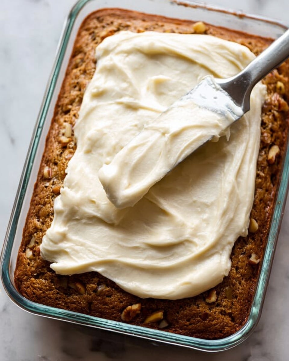 A rectangular glass baking dish holds a single thick layer of brown cake with visible small nut pieces throughout. On top of this cake layer, a thick spread of smooth, creamy white frosting is being evenly moved with a silver spatula. The background surface is a white marbled texture. Photo taken with an iphone --ar 4:5 --v 7