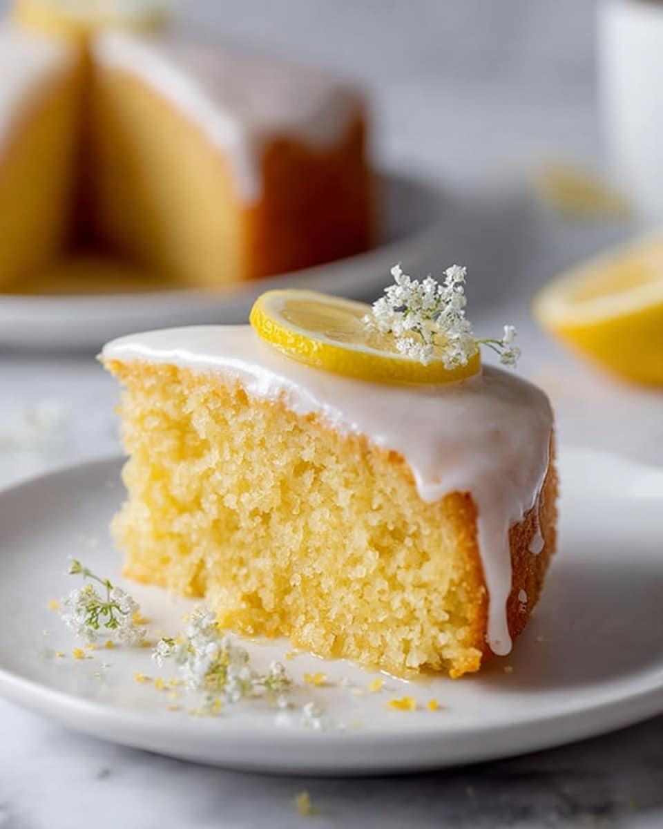 A single slice of yellow cake sits on a white plate placed on a white marbled surface, showing a moist, crumbly texture. The cake has one layer topped with smooth white icing that drips slightly down the sides. On top of the icing, there is a thin, round slice of lemon and a small cluster of tiny white flowers. Some scattered crumbs and a few more small white flowers are placed near the base of the cake on the plate. In the blurred background, another slice of the same cake is visible. Photo taken with an iphone --ar 4:5 --v 7