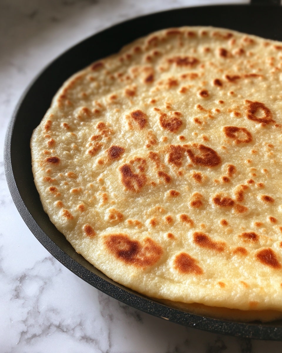 A single round, lightly browned flatbread with a soft texture and small spots of darker tan is cooking on a black pan. The flatbread is slightly puffed up, showing gentle bubbles and a firm yet airy surface. The pan contrasts with the flatbread's warm beige color. The background is a white marbled texture. photo taken with an iphone --ar 4:5 --v 7