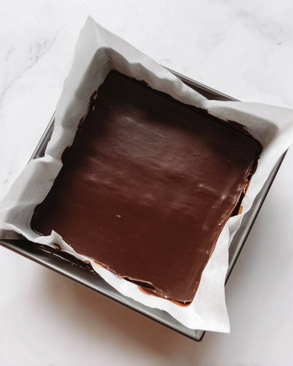 A square pan lined with white baking paper holds a single thick layer of smooth, dark chocolate. The chocolate surface is shiny and even, with no visible cracks or decorations. The white marbled surface beneath adds a clean and bright contrast to the dark chocolate layer. photo taken with an iphone --ar 4:5 --v 7