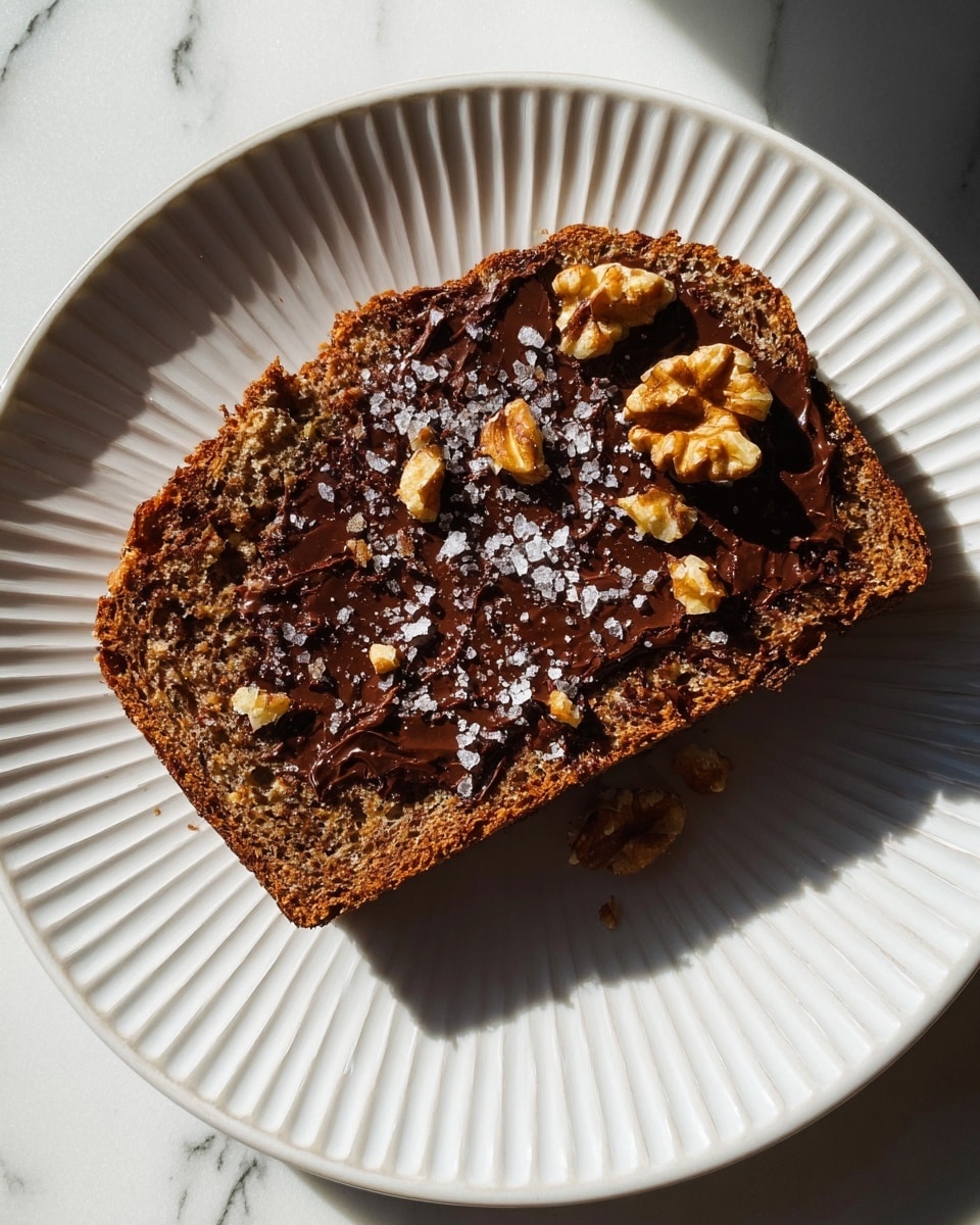 A single thick slice of brown bread with a rough, crumbly texture is placed in the center of a white plate with a ribbed edge. On the bread, a dark, melted chocolate layer is spread unevenly, showing small bits of walnuts peeking through and whole walnut pieces attached to the top right edge. Scattered on the chocolate are small flakes of coarse sea salt, adding a slightly shiny touch. The plate sits on a white marbled surface, with natural light highlighting the textures and shadows around the bread. photo taken with an iphone --ar 4:5 --v 7