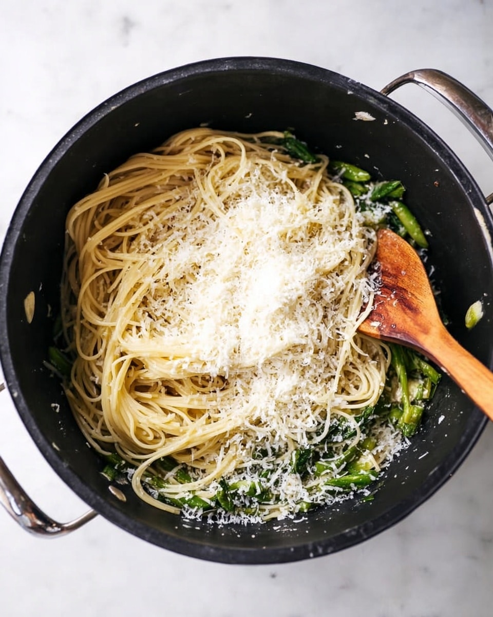 Inside a large black pot, there are three layers visible: at the bottom, bright green cooked vegetables; in the middle, a pile of light creamy yellow spaghetti noodles tangled together; on top, a generous dusting of finely grated white cheese covering part of the noodles and vegetables. A wooden spoon is resting on the right side of the noodles, slightly stirring them. The pot is on a white marbled surface. Photo taken with an iphone --ar 4:5 --v 7
