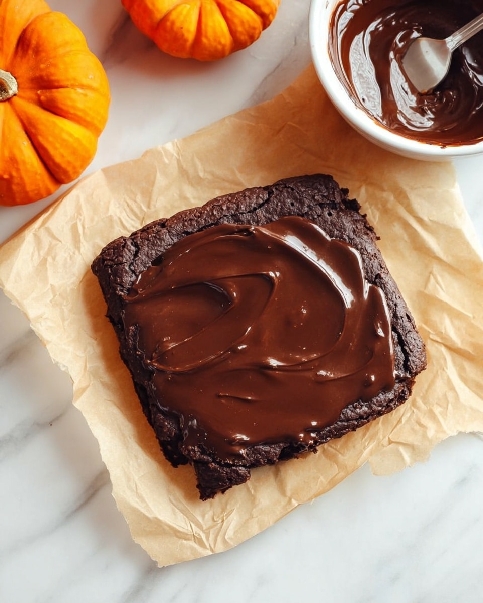 A single square-shaped dark brown brownie sits on a piece of lightly crumpled beige parchment paper on a white marbled surface. The brownie is thick with a slightly rough texture on the edges. A smooth layer of glossy chocolate sauce is spread unevenly on top, with a few visible swirls and reflections. To the side, there is a white bowl filled with more shiny chocolate sauce and a spoon inside it. Two small orange pumpkins are also visible in the upper left corner. Photo taken with an iphone --ar 4:5 --v 7