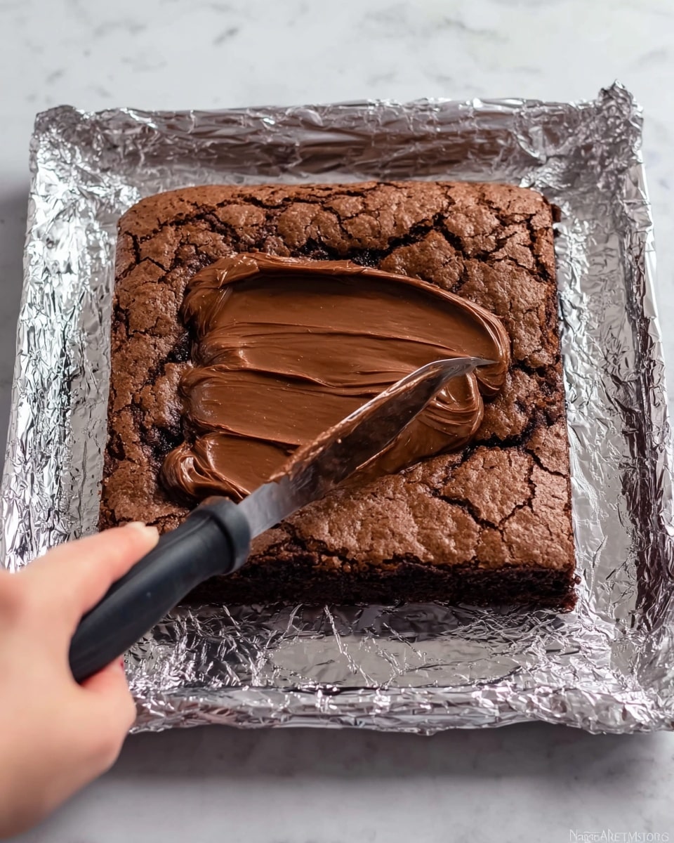 A square chocolate brownie with a rough, cracked top layer rests on shiny aluminum foil over a white marbled surface. A knife with a black handle is spreading smooth, dark chocolate frosting in the center of the brownie, creating a glossy thick layer that contrasts with the matte, textured brownie base. A woman's hand holds the knife from the left side, moving the rich chocolate spread evenly. The scene is bright and clear, showing the details of the brownie crust and the shiny frosting inside the square cut area photo taken with an iphone --ar 4:5 --v 7