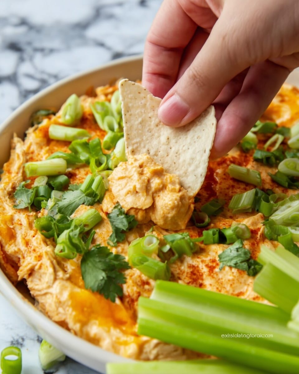 A close-up image shows a woman's hand dipping a beige tortilla chip into a cheesy orange dip spread evenly on a white plate, topped with chopped green onions, fresh cilantro leaves, and a few green celery sticks arranged around the dip, all set on a white marbled surface. Photo taken with an iphone --ar 4:5 --v 7