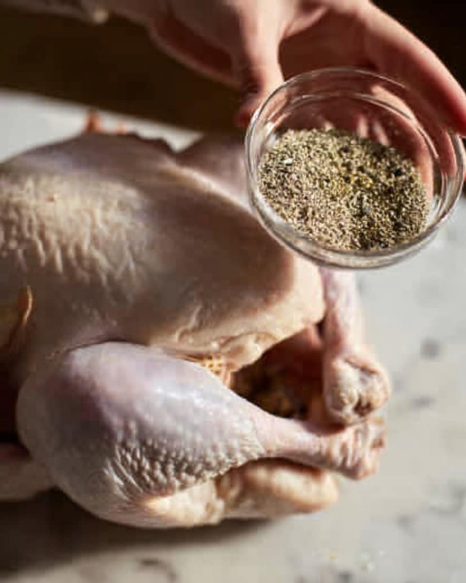 A raw whole chicken lies on a white marbled surface, its pale skin showing natural textures and folds. A woman's hand holds a small clear glass bowl filled with a mix of coarse spices, mostly light green and black seeds, above the chicken's leg, about to sprinkle them. The scene focuses on the chicken's legs and the woman's hand, with a soft and natural light highlighting the textures of the chicken skin and spices. Photo taken with an iphone --ar 4:5 --v 7