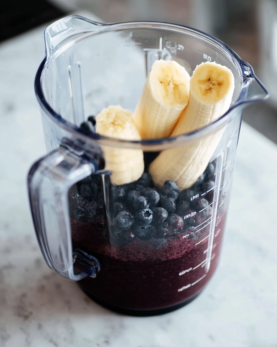 The image shows a clear blender jar filled with two main layers of ingredients: the bottom layer is dark purple blueberry mixture, and the top layer consists of two peeled banana halves that are light yellow with soft texture leaning inside the blender. The blender is placed on a white marbled surface, and the jar has measurement markings on the side. The background is softly blurred, focusing attention on the fresh fruit inside the blender. photo taken with an iphone --ar 4:5 --v 7