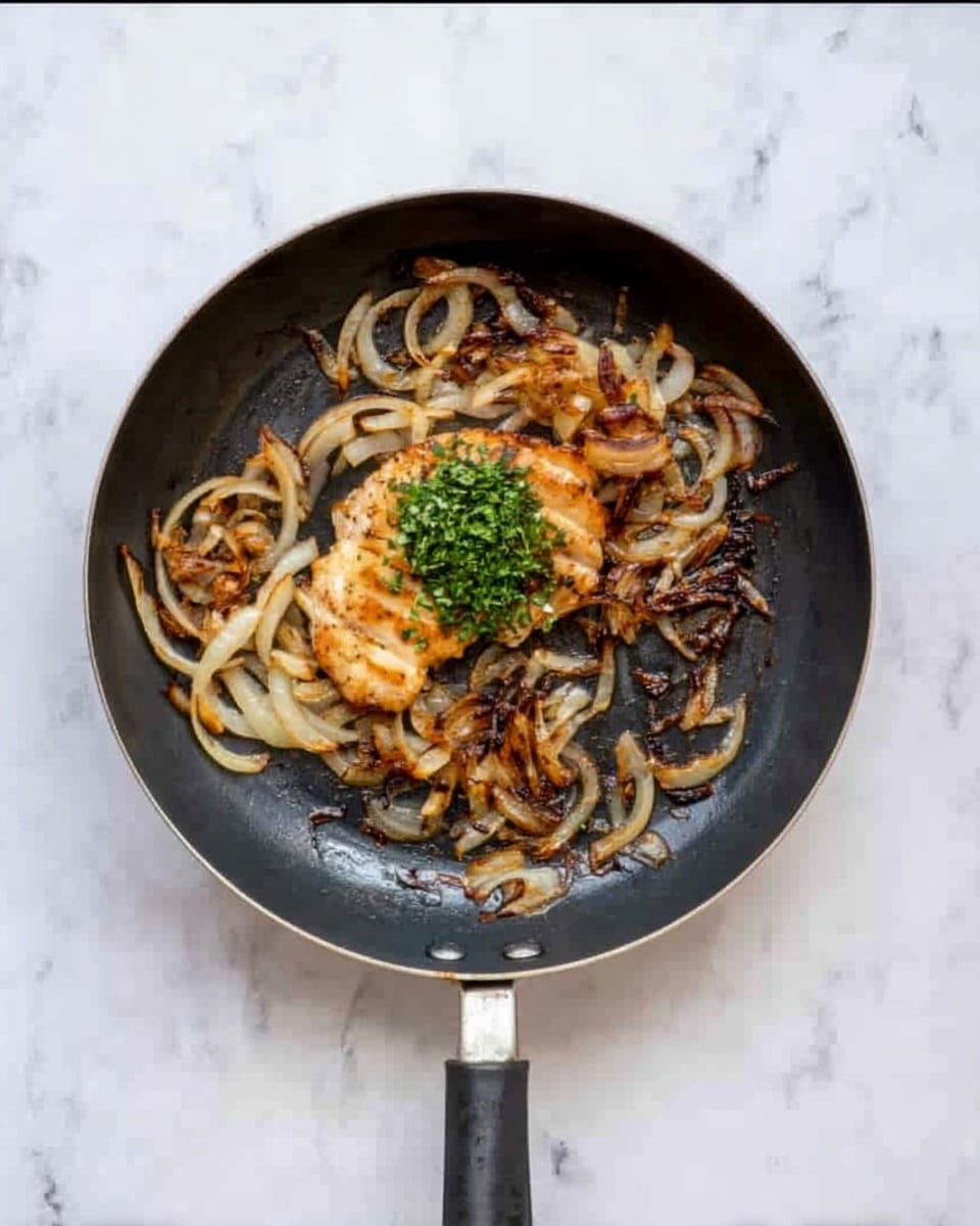 The image shows a black frying pan resting on a white marbled surface with cooked sliced onions and a piece of golden-brown cooked chicken in the center. The pan contains an uneven layer of caramelized onions with a small bunch of chopped green herbs placed near the middle. The chicken piece has a slightly crispy texture with some darker roasted spots. The frying pan handle is visible at the bottom of the image, pointing downward. Photo taken with an iphone --ar 4:5 --v 7