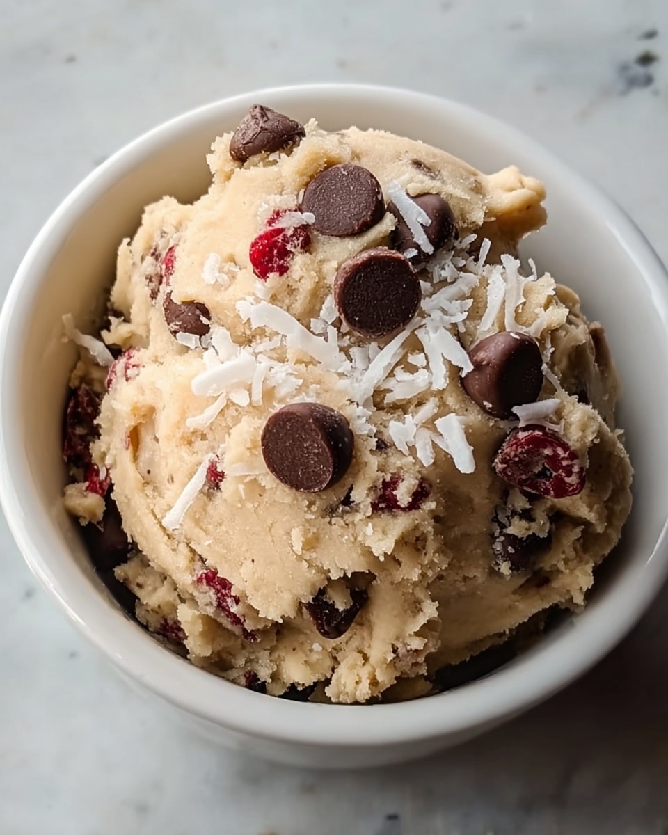 A close-up image of a single scoop of light beige cookie dough in a white bowl. The dough is textured with visible chocolate chips and red dried fruit chunks mixed inside. On top, there are scattered larger chocolate chips and shredded white coconut flakes. The bowl sits on a white marbled surface, providing a clean contrast. The dough looks soft and slightly crumbly with a natural homemade feel. photo taken with an iphone --ar 4:5 --v 7