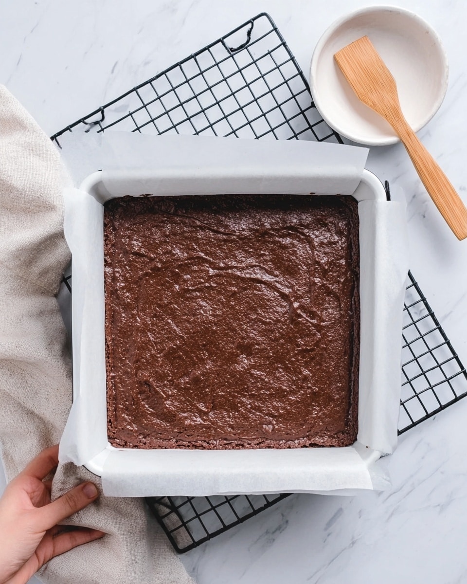 The image shows a white square pan lined with white parchment paper, filled with one thick layer of smooth, rich chocolate brownie batter that has a slightly cracked and uneven texture on top. The pan is placed on a white marbled surface with a grid cloth underneath it. Above and to the right side, a woman’s hand is resting on a small white dish with a wooden spatula on it. photo taken with an iphone --ar 4:5 --v 7