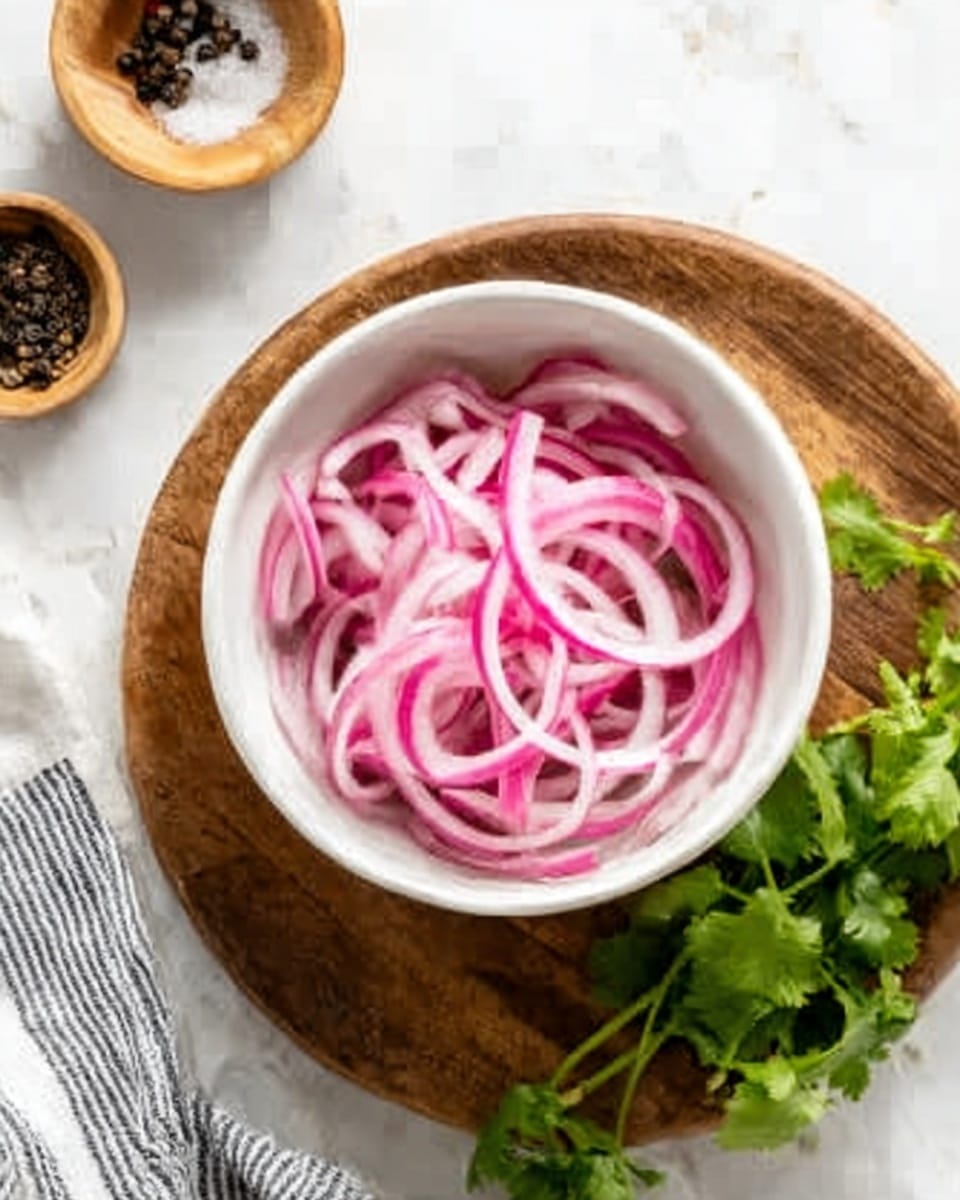 A small white bowl filled with thin, curly slices of pink and white pickled onions sits on a round wooden board. The onions have a slightly shiny, wet texture and are loosely piled inside the bowl. To the right side of the board, fresh green cilantro leaves rest partly on a white marbled surface with a striped cloth nearby. Above the bowl, two small light wooden bowls hold black pepper and salt. The scene is bright with natural light, showing clear details and soft shadows. Photo taken with an iphone --ar 4:5 --v 7