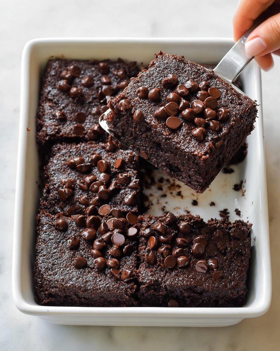 A square white dish holds four large, thick brownies with a rich dark brown texture and a moist, slightly crumbly look. The tops of the brownies are covered with many small, round, shiny chocolate chips, adding a glossy contrast. One brownie is lifted by a woman's hand using a metal spatula, showing its soft and dense inside. The white marbled surface beneath the dish reflects soft light, highlighting the dark chocolate colors. photo taken with an iphone --ar 4:5 --v 7
