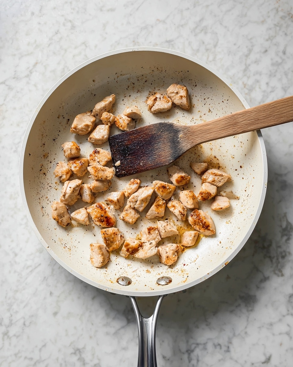 The image shows a white frying pan placed on a white marbled surface. Inside the pan, there are small pieces of browned chicken scattered evenly across the pan's surface. The chicken pieces are light golden with some darker brown spots indicating cooking. A wooden spatula with a darkened and worn texture rests inside the pan, touching some of the chicken pieces. The pan has a shiny metal handle extending upwards from the frame. photo taken with an iphone --ar 4:5 --v 7