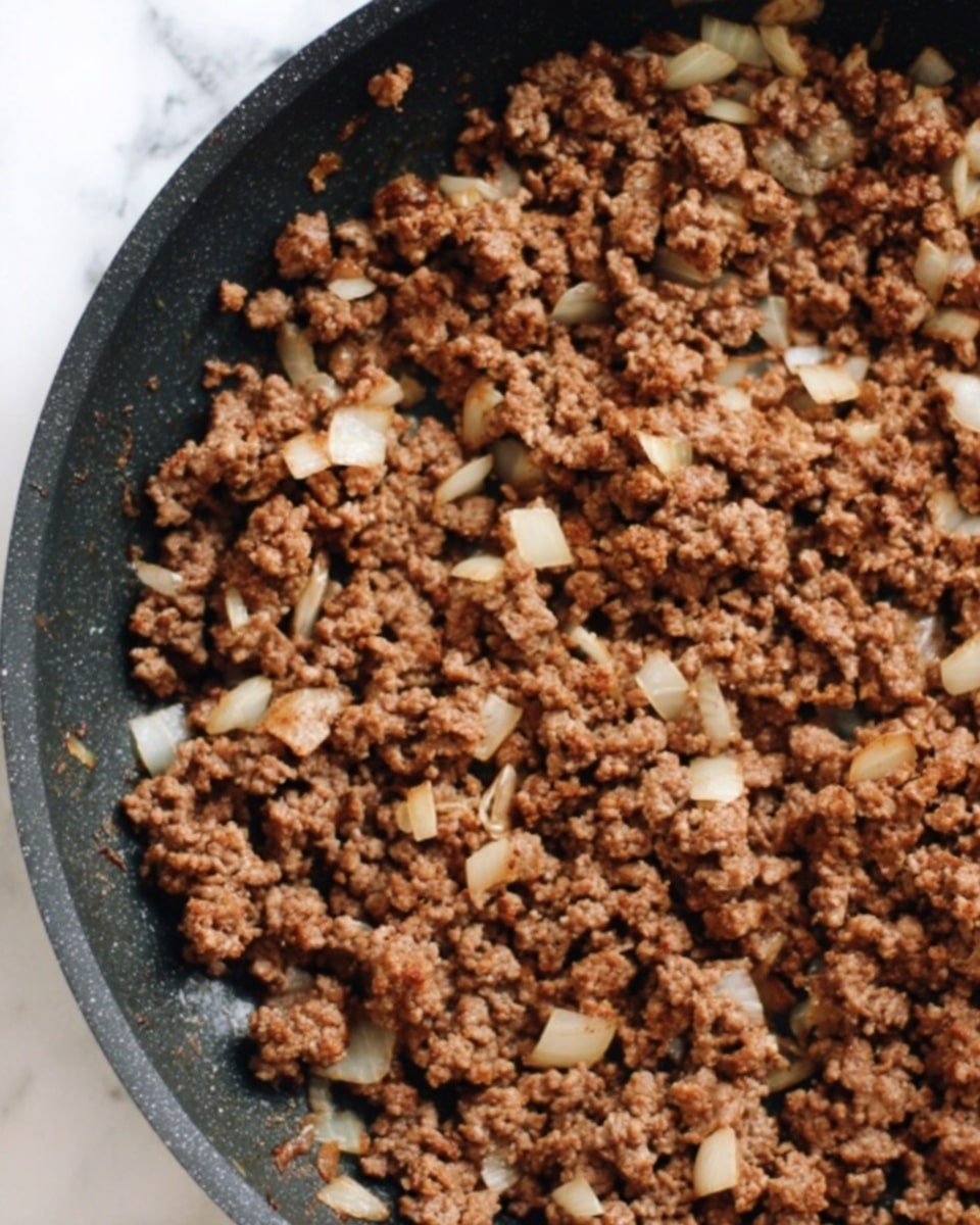 The image shows a close-up view of a black frying pan filled with browned ground meat mixed with small pieces of light-colored onion, all cooked and evenly spread across the pan's surface. The texture looks crumbly with bits of onion adding some lighter patches among the rich brown color of the meat, against a white marbled background. Photo taken with an iphone --ar 4:5 --v 7