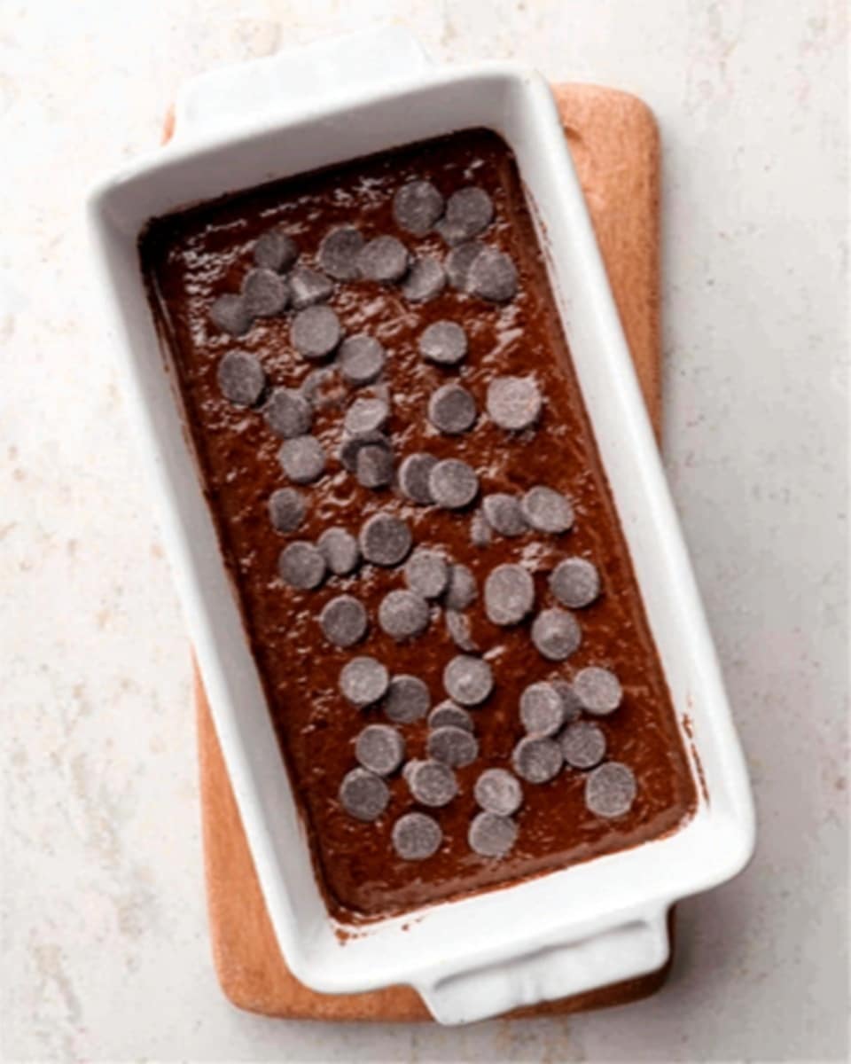 The image shows a white rectangular baking dish with a chocolate batter inside. The batter is dark brown and smooth, filling the dish almost to the top. On the surface, there are many dark chocolate chips scattered evenly across, adding texture and visual interest. The dish is placed on a white marbled surface, creating a clean and bright background. photo taken with an iphone --ar 4:5 --v 7