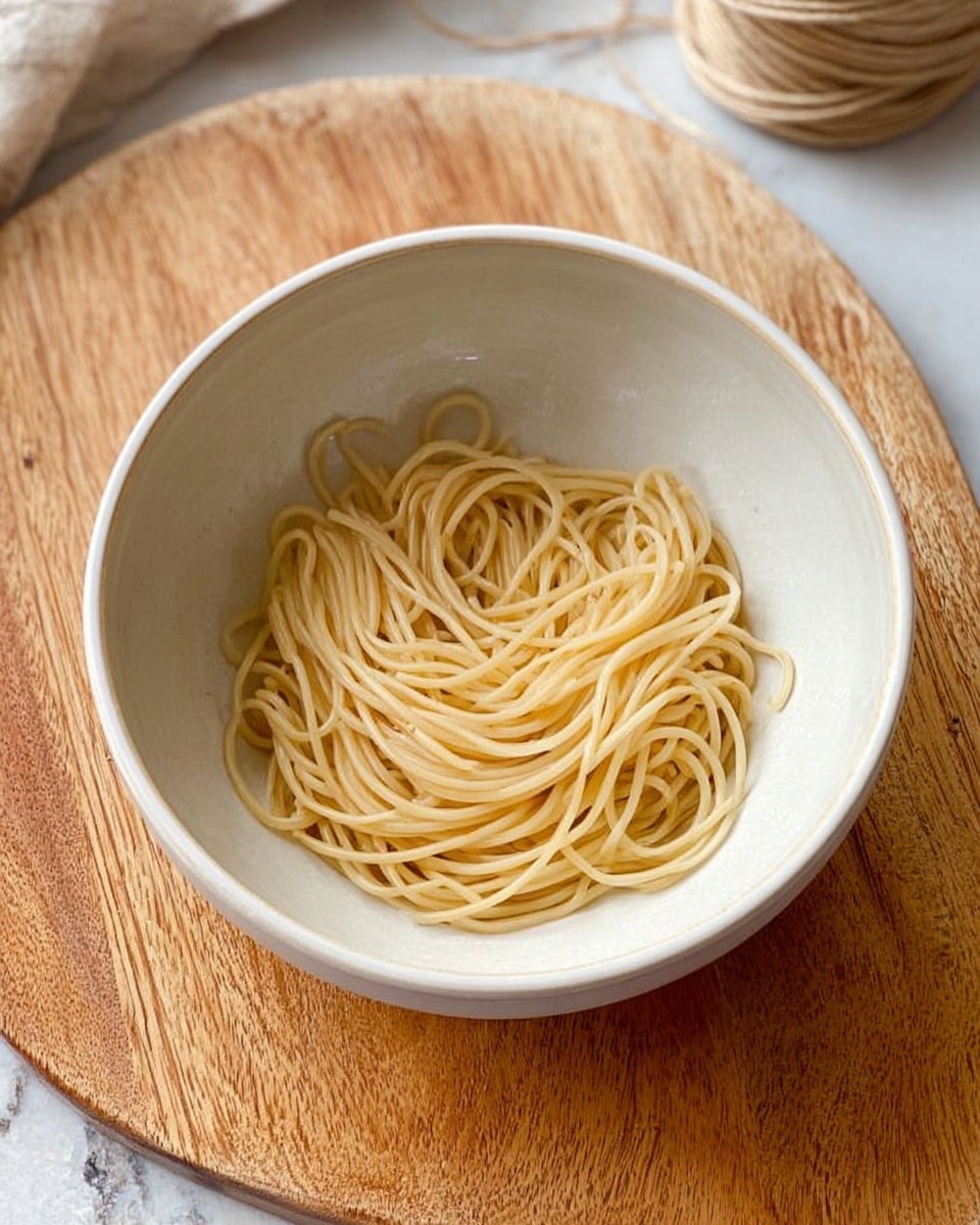 A white bowl sits on a wooden board with thin, light tan strands of noodles loosely coiled inside. The noodles have a smooth, slightly shiny texture and are evenly distributed in the center of the bowl. The background shows part of a white marbled surface, with a spool of string near the top right edge. Photo taken with an iphone --ar 4:5 --v 7