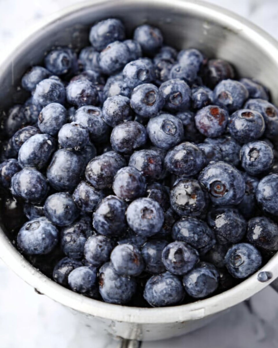 The image shows a close-up of a white bowl filled with fresh blueberries. The small round blueberries are deep blue with a light powdery coating. They fill the bowl fully, with some blueberries near the edges slightly pressed against the bowl. The bowl sits on a white marbled surface, giving a clean and bright background to the rich blue colors of the fruit. Photo taken with an iphone --ar 4:5 --v 7