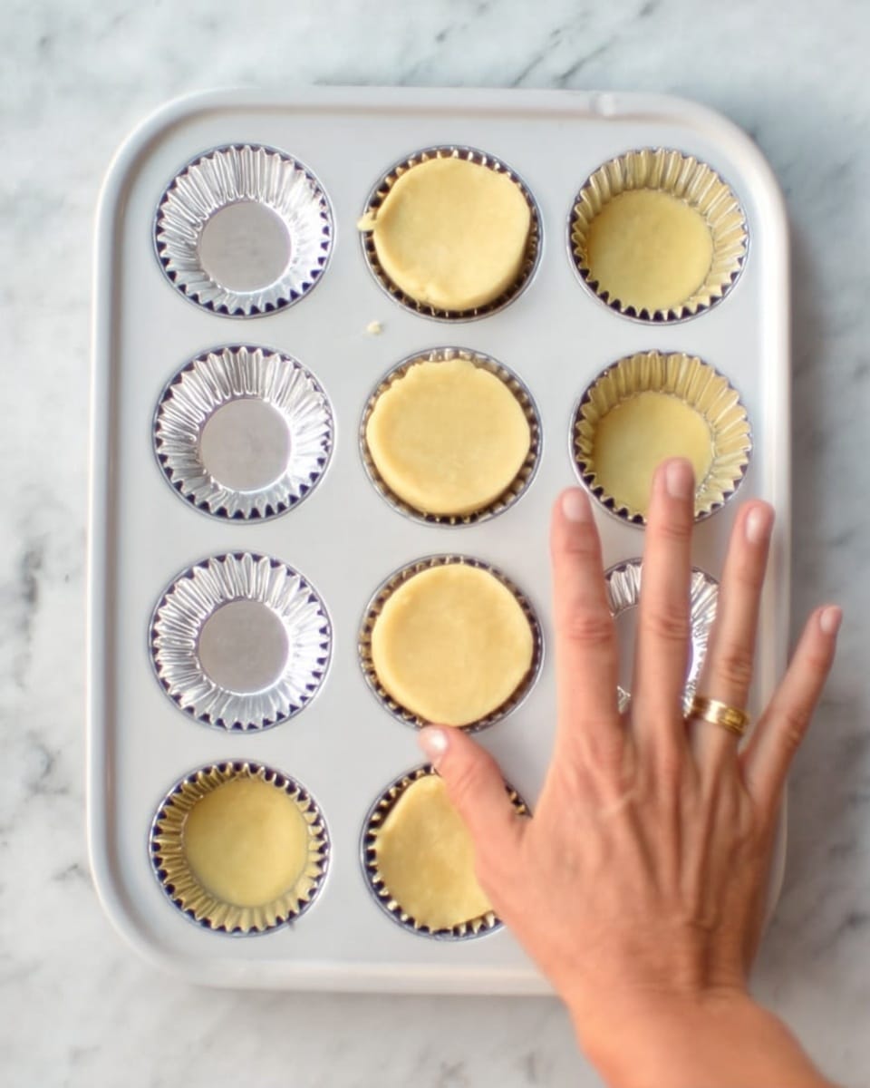 A white metal muffin tray with twelve compartments is placed on a white marbled surface. Six of the compartments hold silver foil cupcake liners, and the other six contain round, even layers of light yellow dough pressed neatly inside. A woman’s hand with a gold ring is pressing the dough into one of the compartments, shaping the edges to fit the liner. The scene shows the process of lining the muffin tray with dough before baking. Photo taken with an iphone --ar 4:5 --v 7