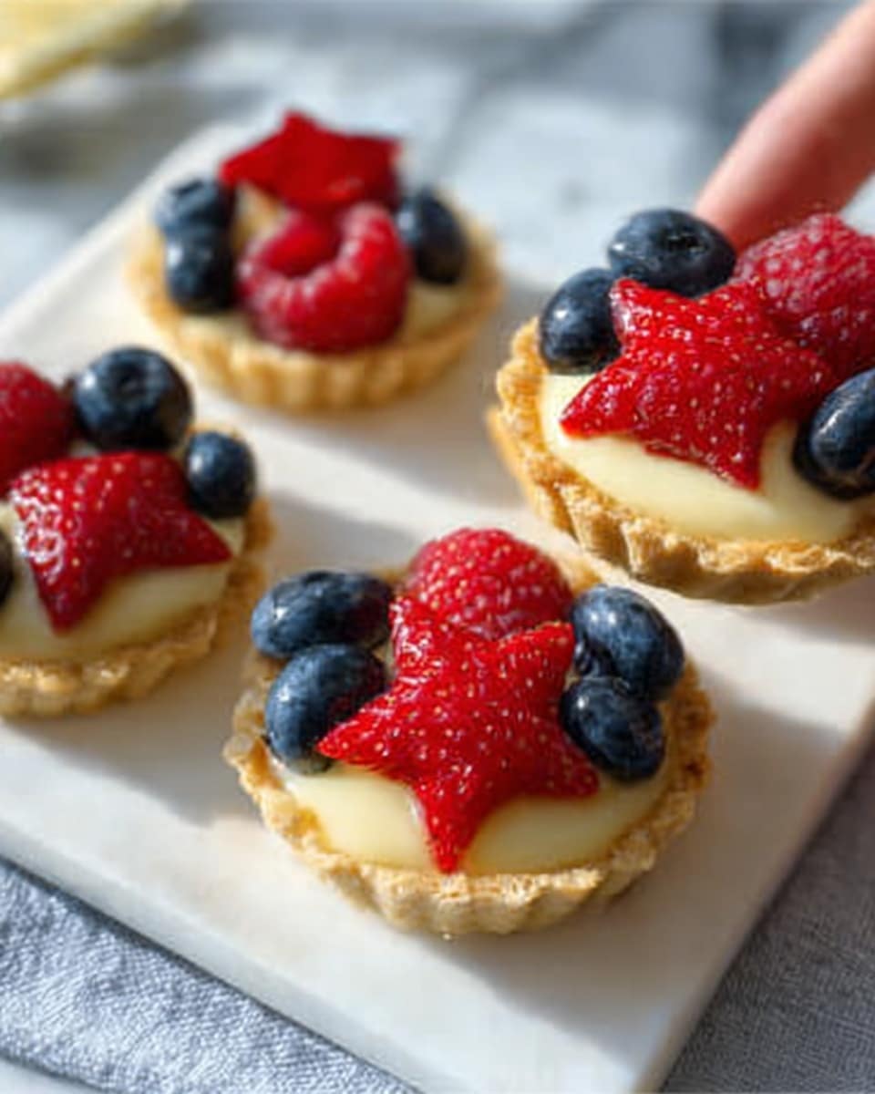 The image shows small tartlets on a white square plate with a white marbled surface underneath. Each tartlet has a golden-brown, crispy crust filled with smooth creamy custard. On top of the custard, there are layers of fresh fruit: deep red strawberries cut into star shapes and bright red round raspberries, paired with clusters of shiny dark blue blueberries. The fruits are arranged neatly, with the star-shaped strawberries standing out on each tartlet. A woman's hand holds one tartlet, lifting it slightly from the plate. The light is soft and natural, highlighting the glossy textures of the fruit and the creamy filling. Photo taken with an iphone --ar 4:5 --v 7