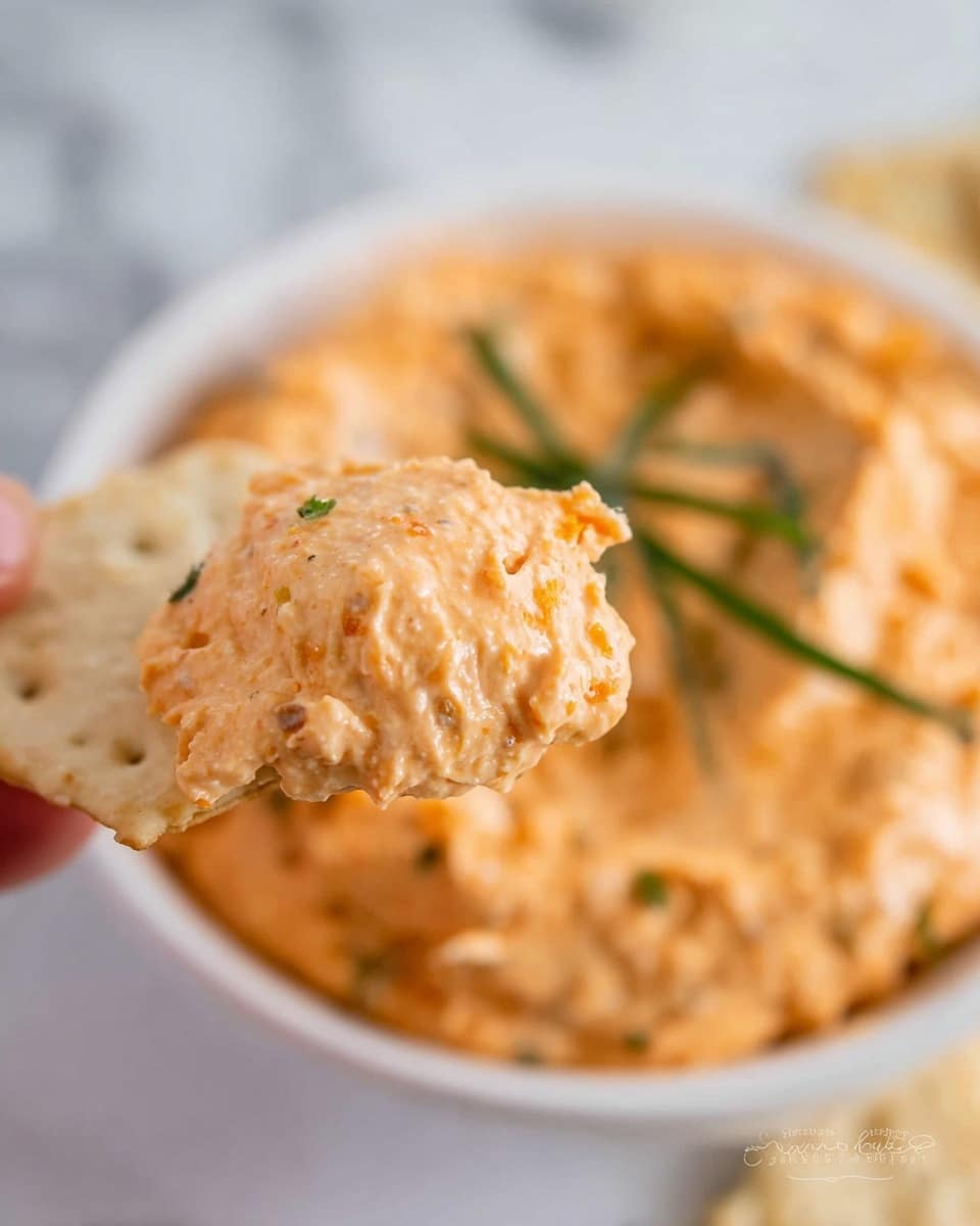 A close-up shows a white cracker held by a woman's hand, dipped in a thick, creamy orange spread with visible texture and small bits. The spread is layered on the cracker in a soft, uneven way. Below, a white bowl full of the same orange spread with a few sprigs of green herbs sprinkled on top is placed on a white marbled surface. The image focuses on the cracker and spread, with the bowl blurred in the background. photo taken with an iphone --ar 4:5 --v 7