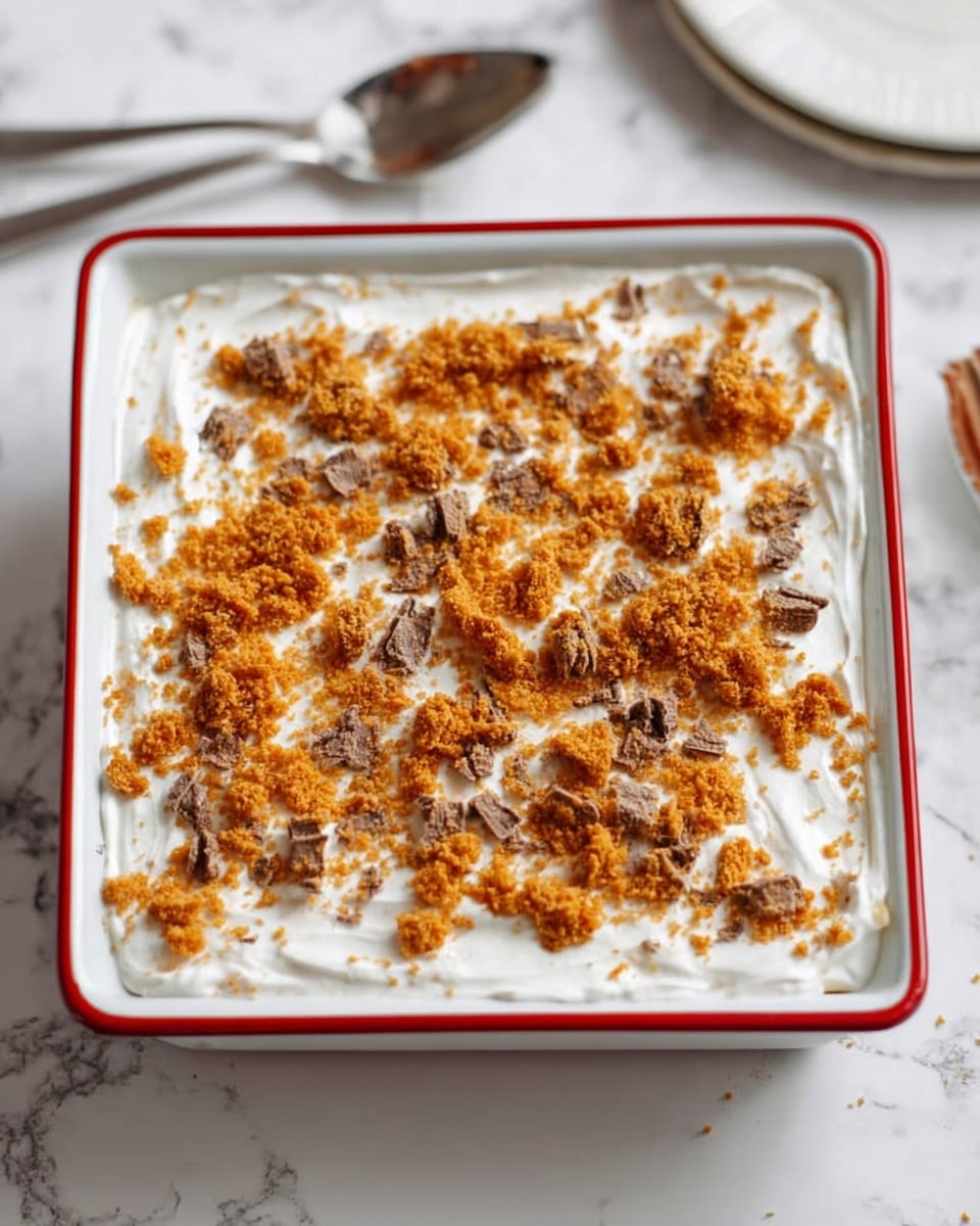 A square white dish with a red rim holds a dessert with two visible layers. The bottom layer is smooth and white, topped with a thick layer of white cream that has a soft, slightly textured surface. On top, there are scattered small orange crumb pieces mixed with bits of brown chocolate, spread evenly across the cream. The dish sits on a white marbled surface with a silver utensil placed nearby. photo taken with an iphone --ar 4:5 --v 7