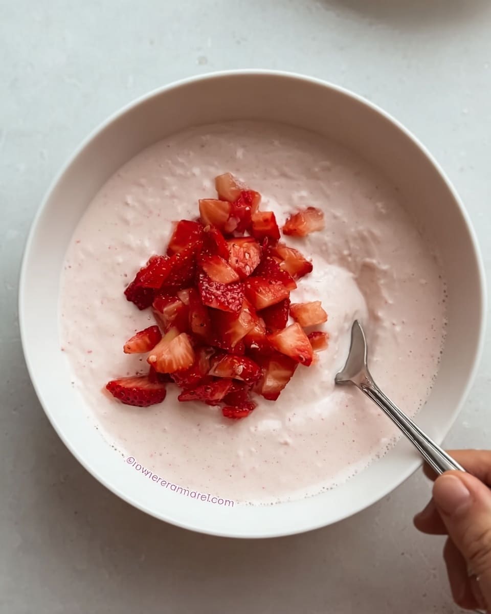 A white bowl filled with a creamy, light pink mixture that has a smooth texture. In the center, there is a small pile of finely chopped strawberries with a bright red color, adding contrast to the pale pink base. A spoon is dipped into the mixture from the right side, partially submerged, with a woman's hand holding it. The bowl is placed on a white marbled surface. photo taken with an iphone --ar 4:5 --v 7