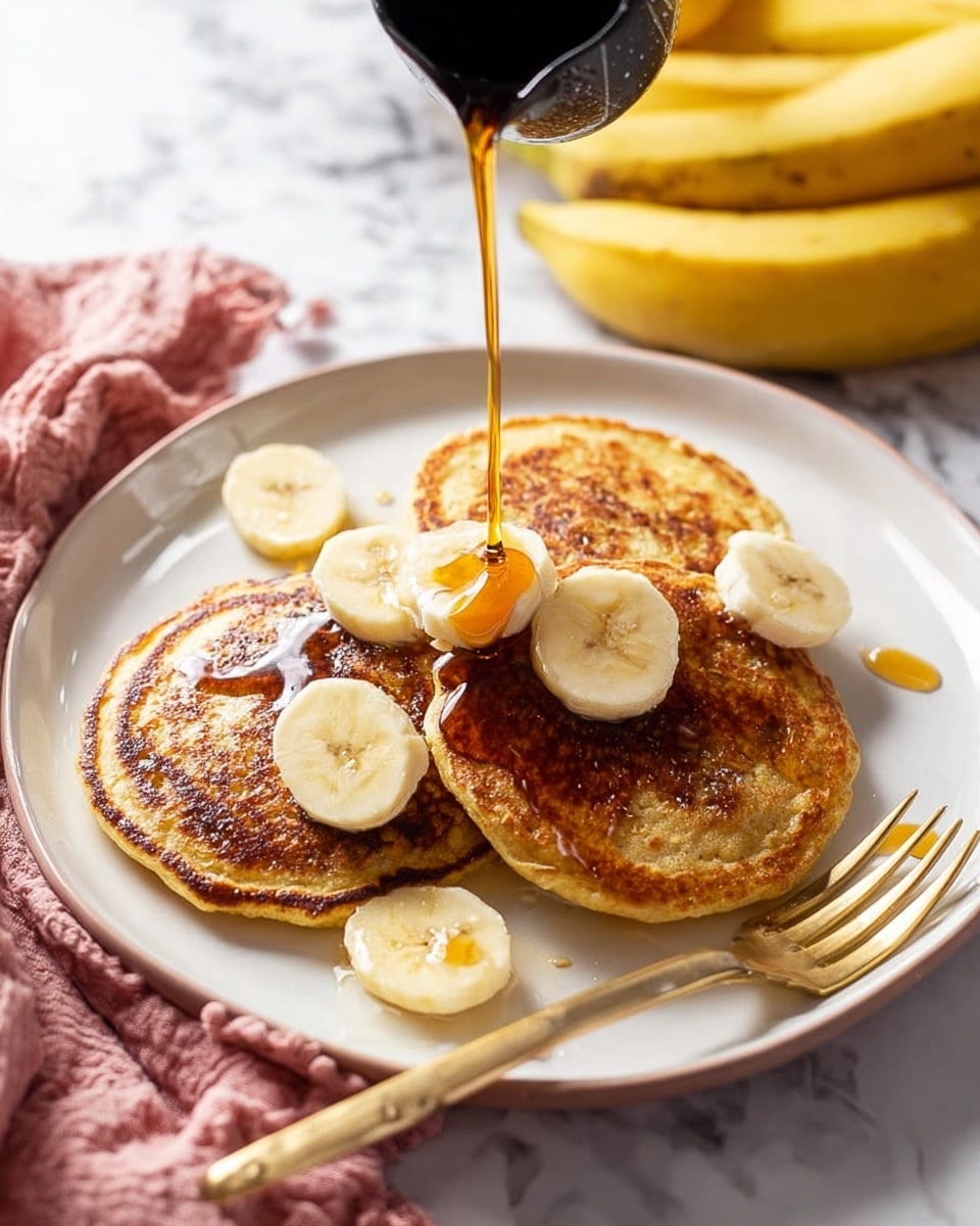 Three round, golden-brown pancakes with a slightly uneven surface are stacked on a white plate. On top and around the pancakes, there are slices of light yellow banana evenly placed. A dark maple syrup is being poured from a black pitcher onto the pancakes, creating shiny reflections and small pools of syrup. A gold fork rests on the right edge of the plate, partly leaning on the pancakes. The plate sits on a white marbled surface with a bunch of bananas and a pink cloth nearby. Photo taken with an iphone --ar 4:5 --v 7