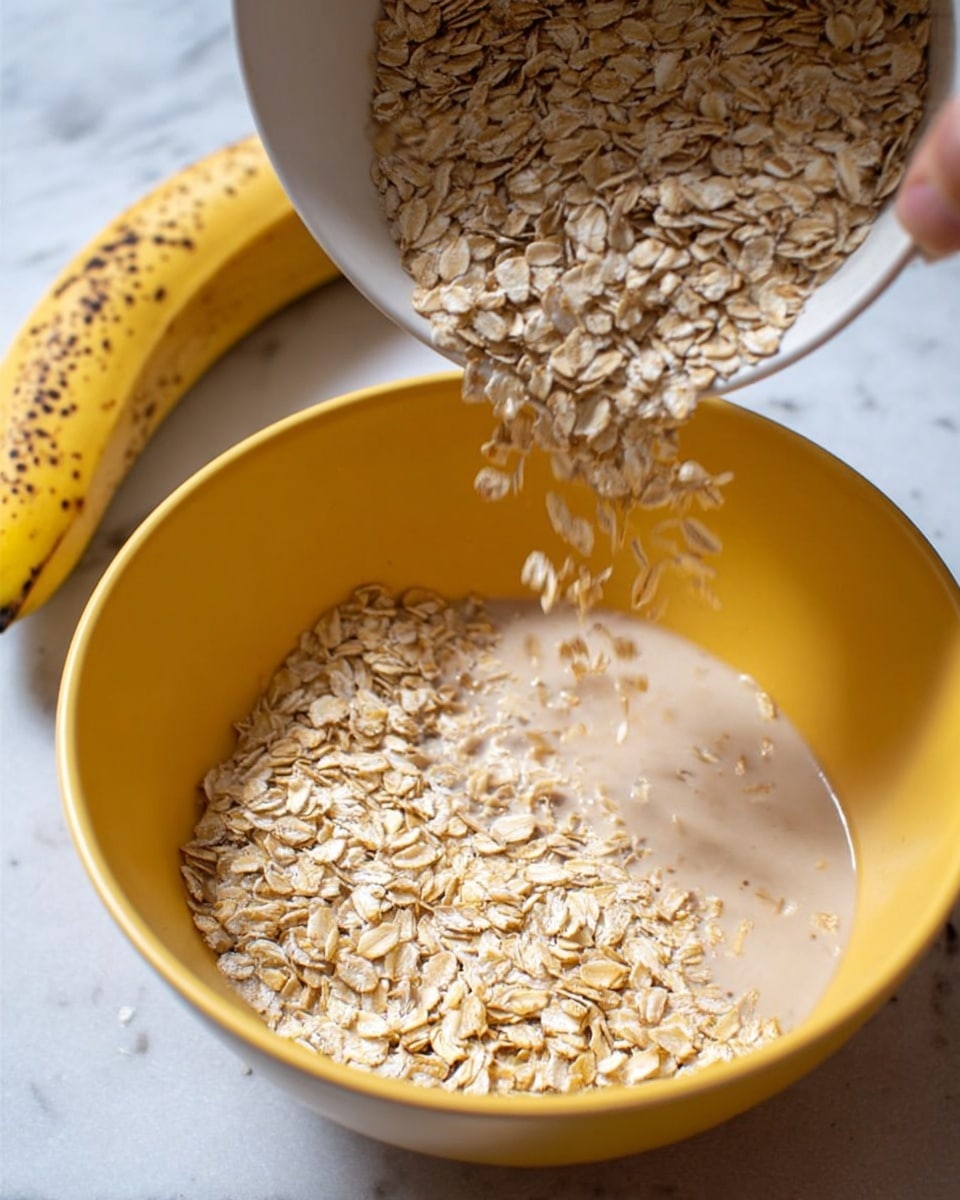 A close-up image shows a yellow bowl filled with a light beige liquid topped with rolled oats. A white bowl, held by a woman's hand at the top left corner, is pouring more rolled oats into the yellow bowl. The texture of the oats is dry and flaky, while the liquid looks smooth and creamy. In the background, there is a ripe banana with spots on its peel, resting on a white marbled surface. The scene is bright and clear, focusing on the action of oats being added to the liquid. Photo taken with an iphone --ar 4:5 --v 7
