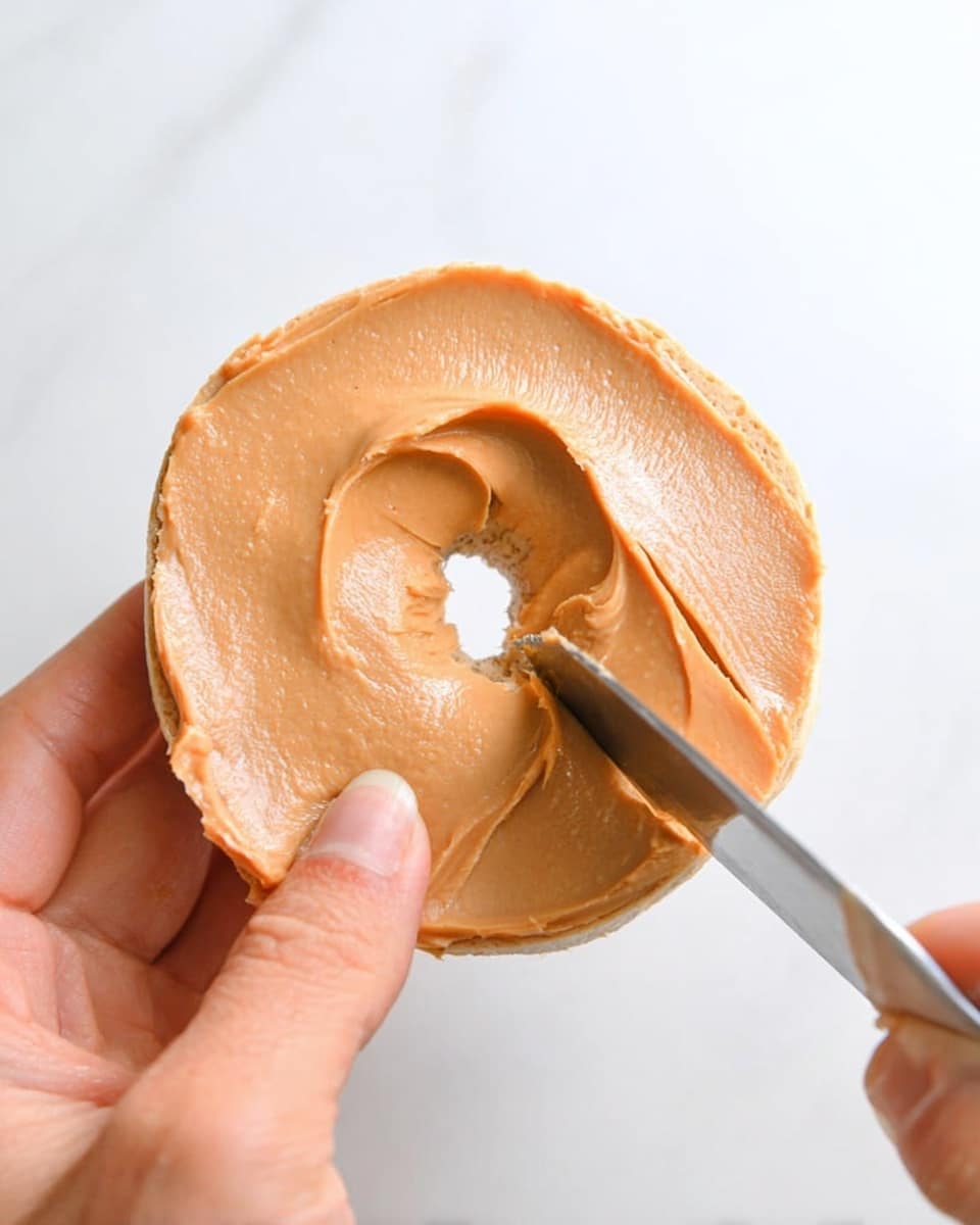A close-up of a woman's hand holding a plain round bagel with a visible hole in the center, spread with a smooth layer of light brown peanut butter. A knife with a smooth edge is applying more peanut butter on the bagel from the bottom right side. The background is white marbled texture, highlighting the smooth creamy texture and light tan color of the peanut butter against the plain bagel skin. Photo taken with an iphone --ar 4:5 --v 7