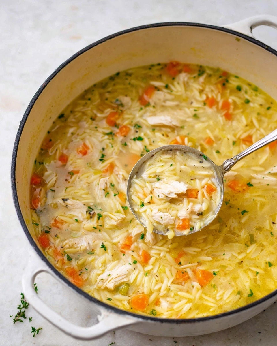 A large white pot filled with a creamy chicken soup with orzo pasta. The soup has layers of tender white chicken pieces, small bright orange carrot cubes, and tiny green herb bits spread evenly in a thick yellowish broth. The orzo pasta looks soft and cooked, covering much of the surface. A silver spoon is partially dipped into the soup, with some broth and ingredients held slightly above the pot’s edge. The pot sits on a white marbled surface with a few small herb pieces scattered around. photo taken with an iphone --ar 4:5 --v 7